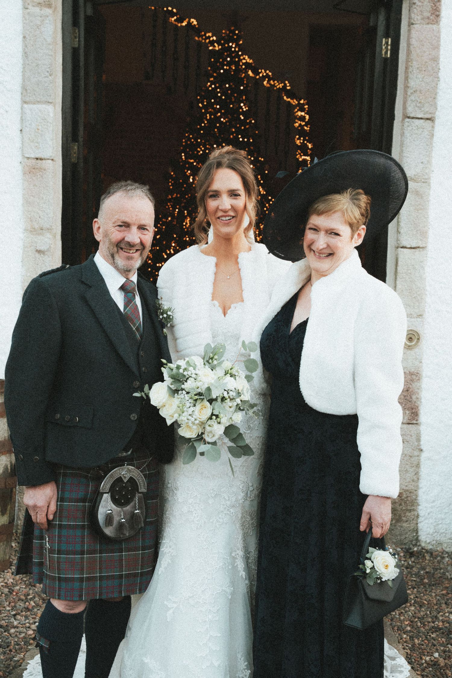 Wedding group poses by white building with kilts and formal winter attire.