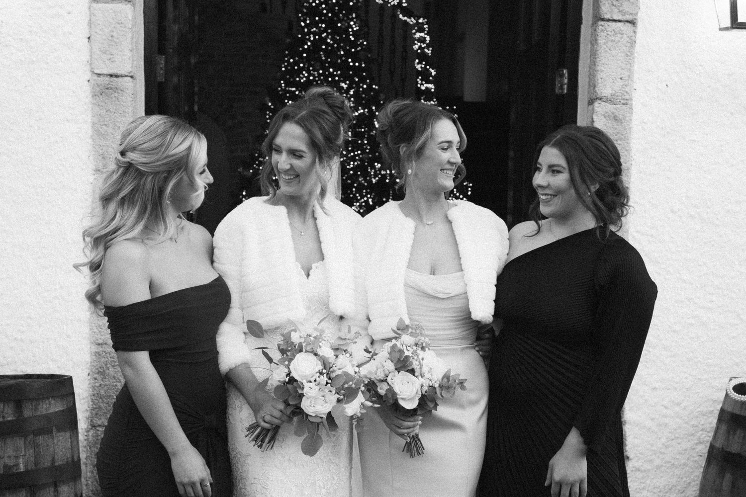 Wedding party members share candid moments outside white stone venue with decorative lighting.