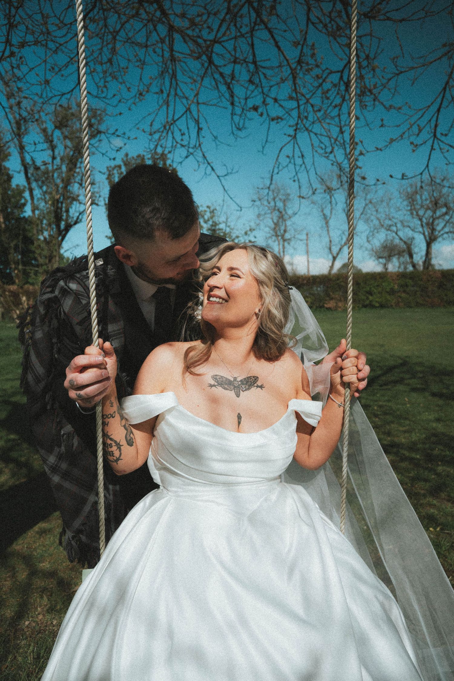Bride in white off-shoulder wedding gown sits on rope swing while groom kisses her cheek outdoors.