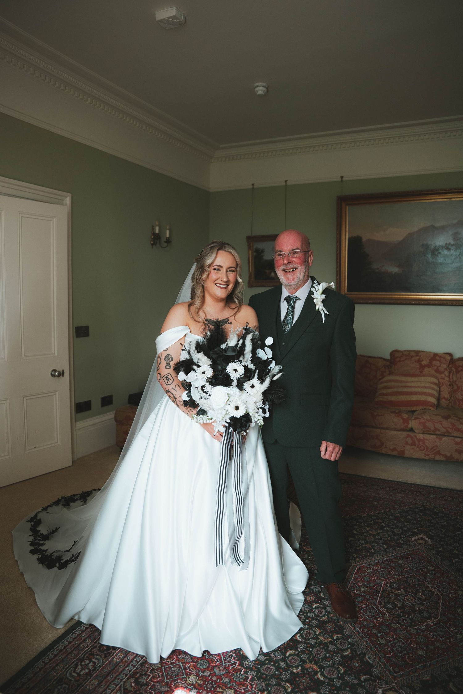 Bride in off-shoulder white gown holding black and white bouquet poses with father in dark suit indoors.