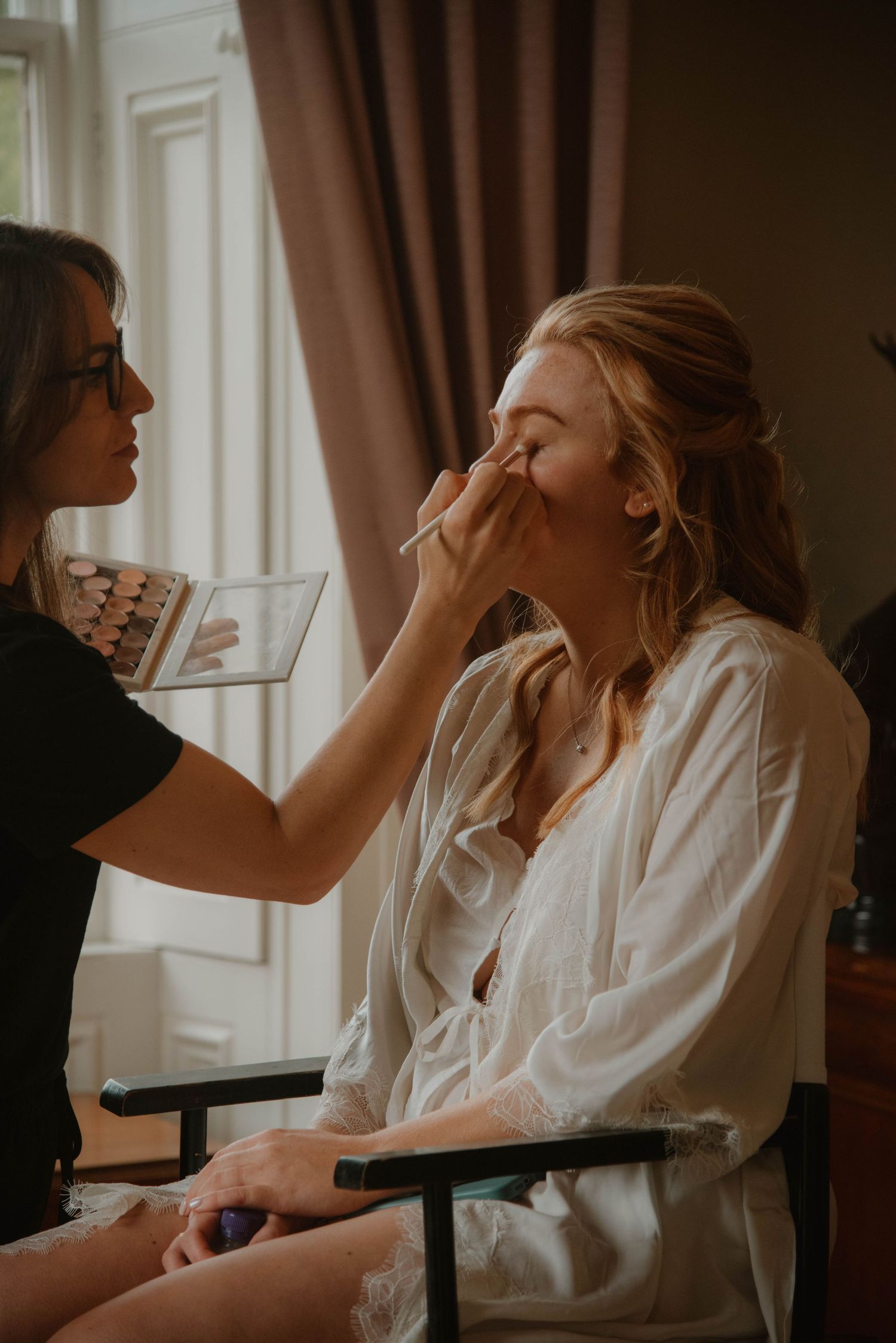 Soft natural light illuminates bridal preparation scene with makeup artist at work.