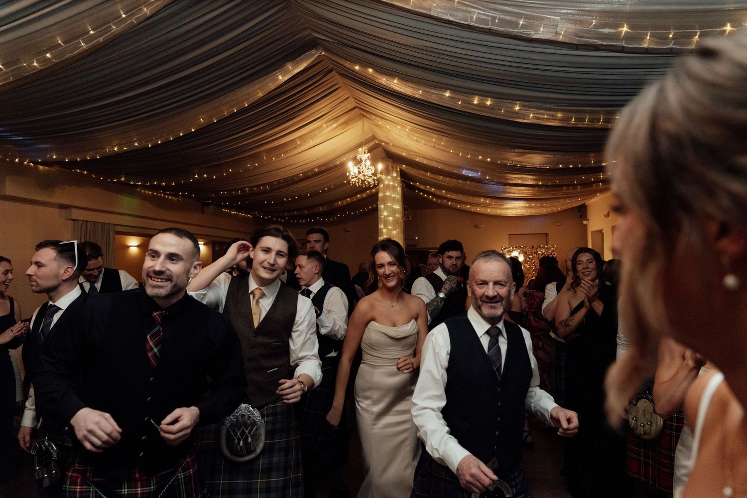 Wedding reception guests dancing and celebrating under draped ceiling with string lights.