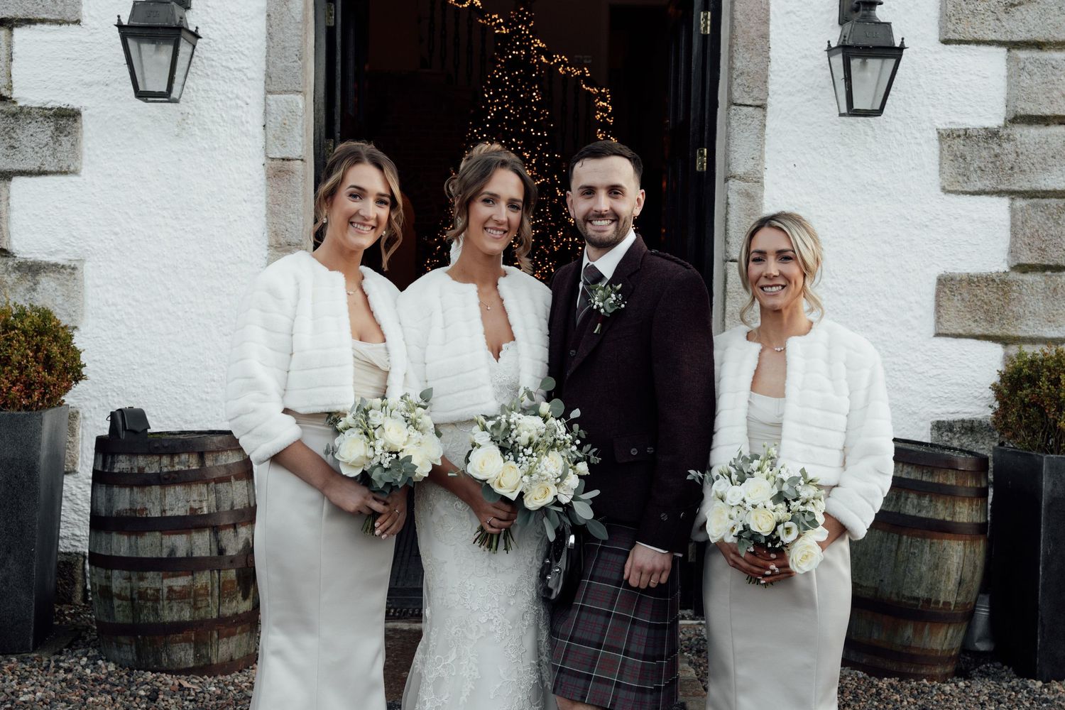 Wedding party poses outside stone building with traditional Scottish kilts and white dresses.