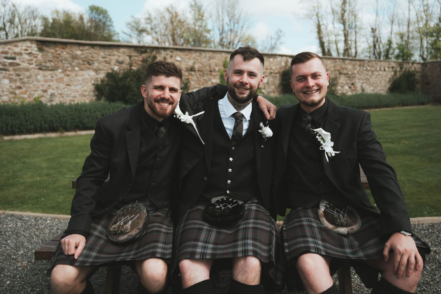 Three men in Scottish kilts and formal jackets smile together on a bench at an outdoor wedding celebration.