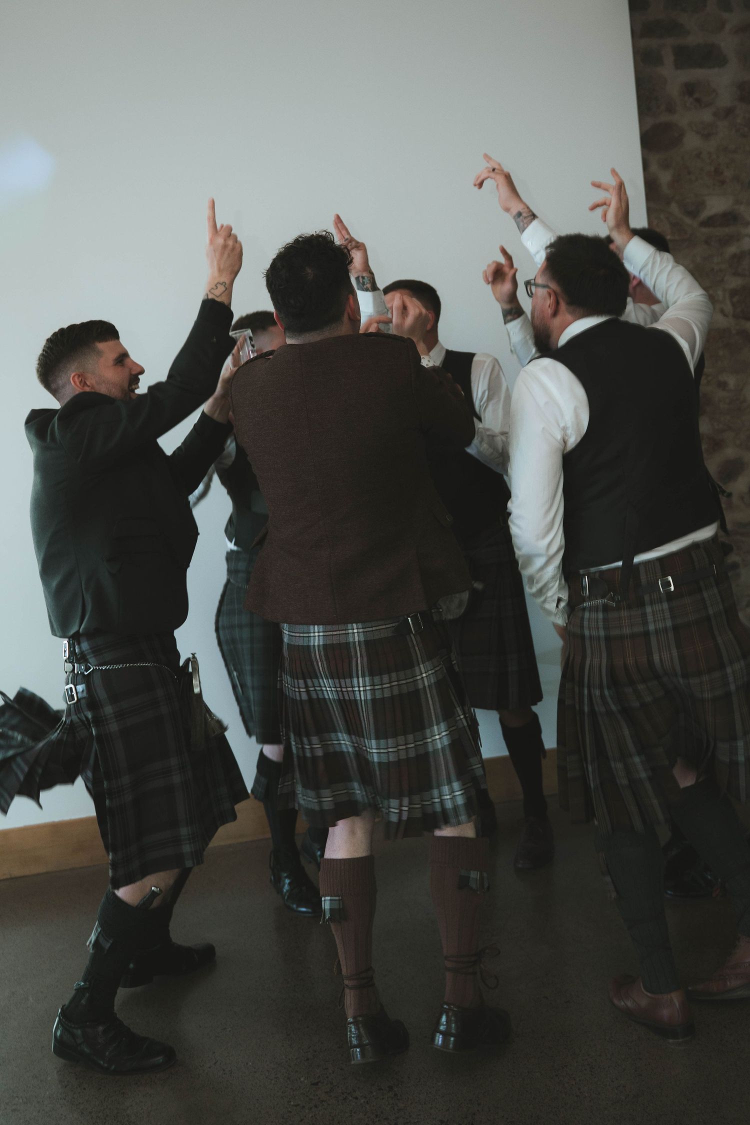 Group of groomsmen in Scottish kilts cheering with arms raised at wedding reception.