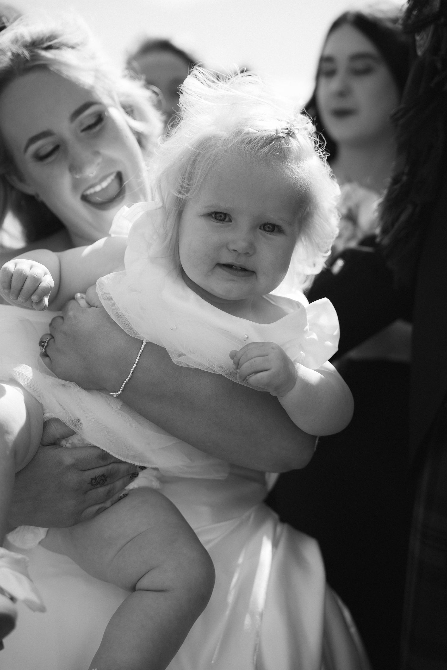 Black and white portrait of a curly-haired baby in white dress being held at a wedding.