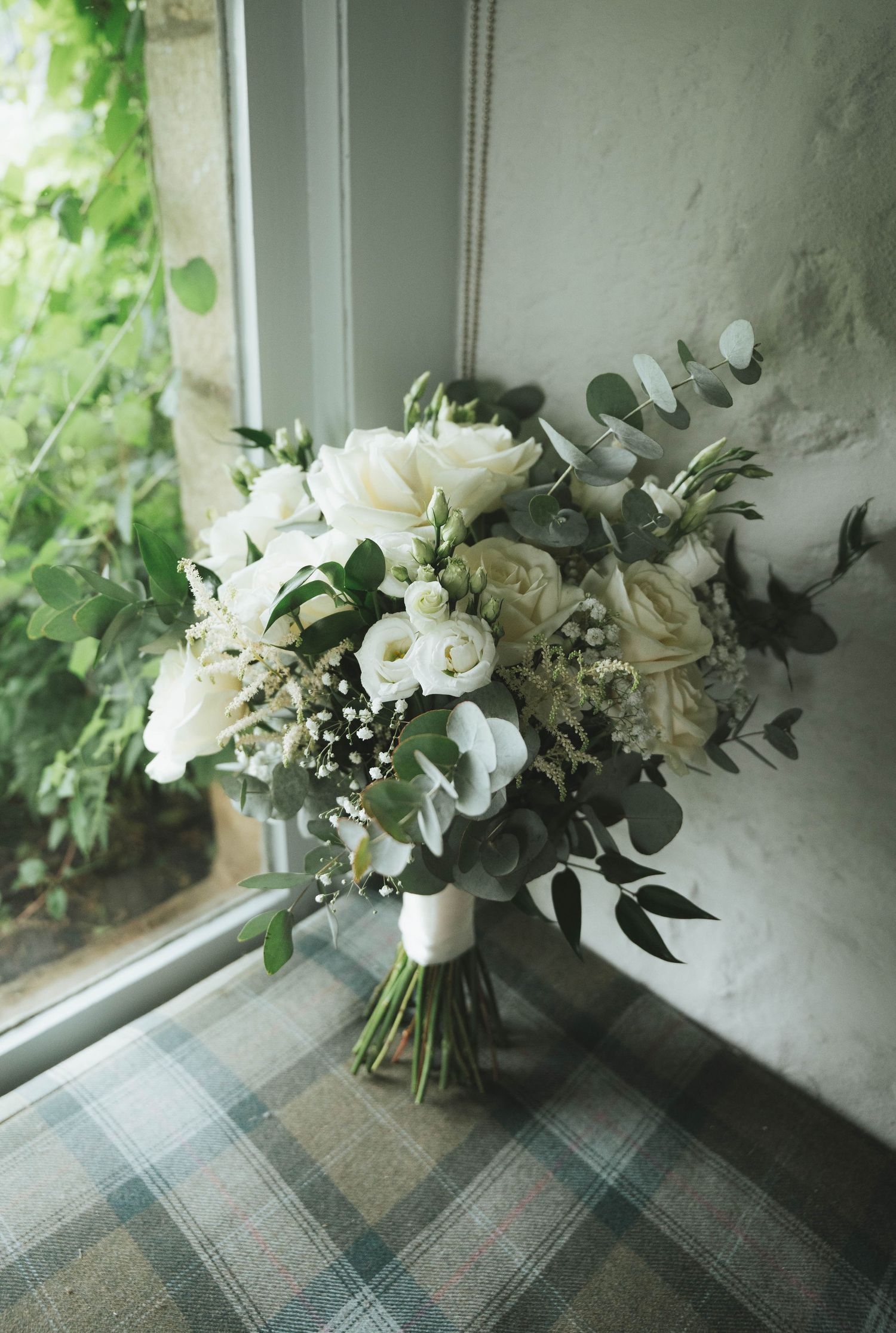 Elegant wedding bouquet of white roses, lisianthus, and eucalyptus resting on a plaid chair by a window.