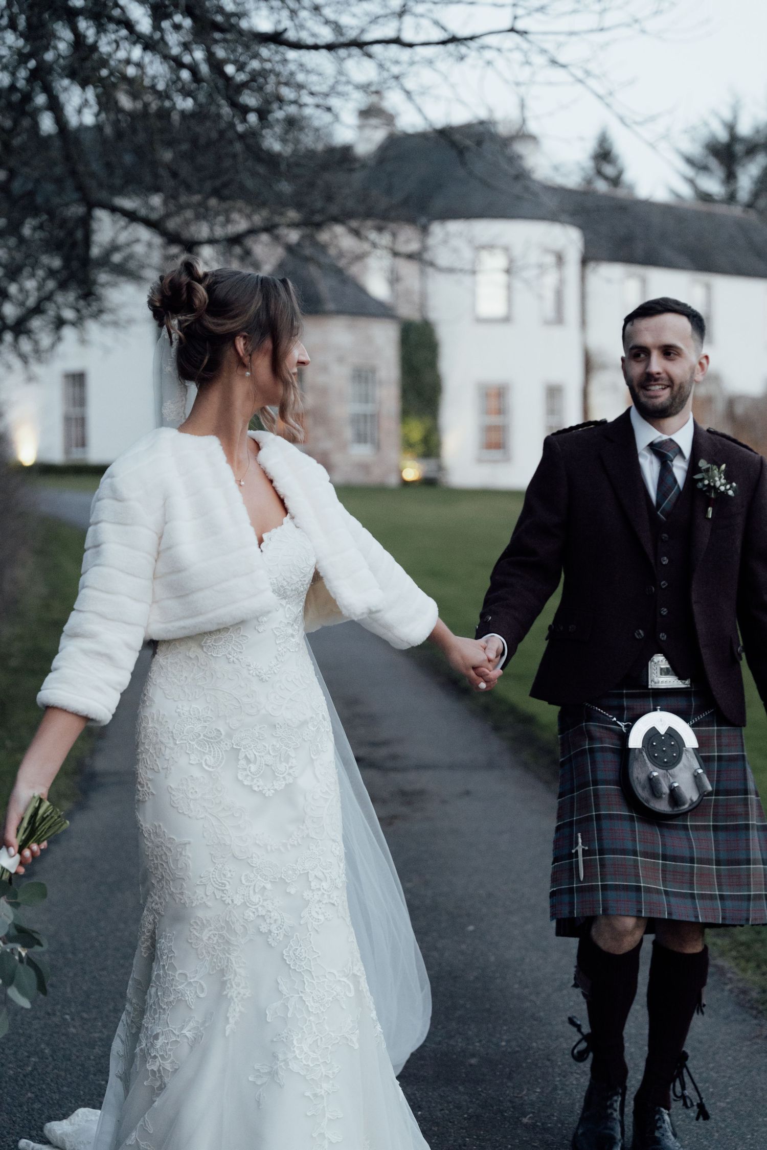 Bride in lace dress and groom in Scottish kilt walk together on a path near a grand estate.