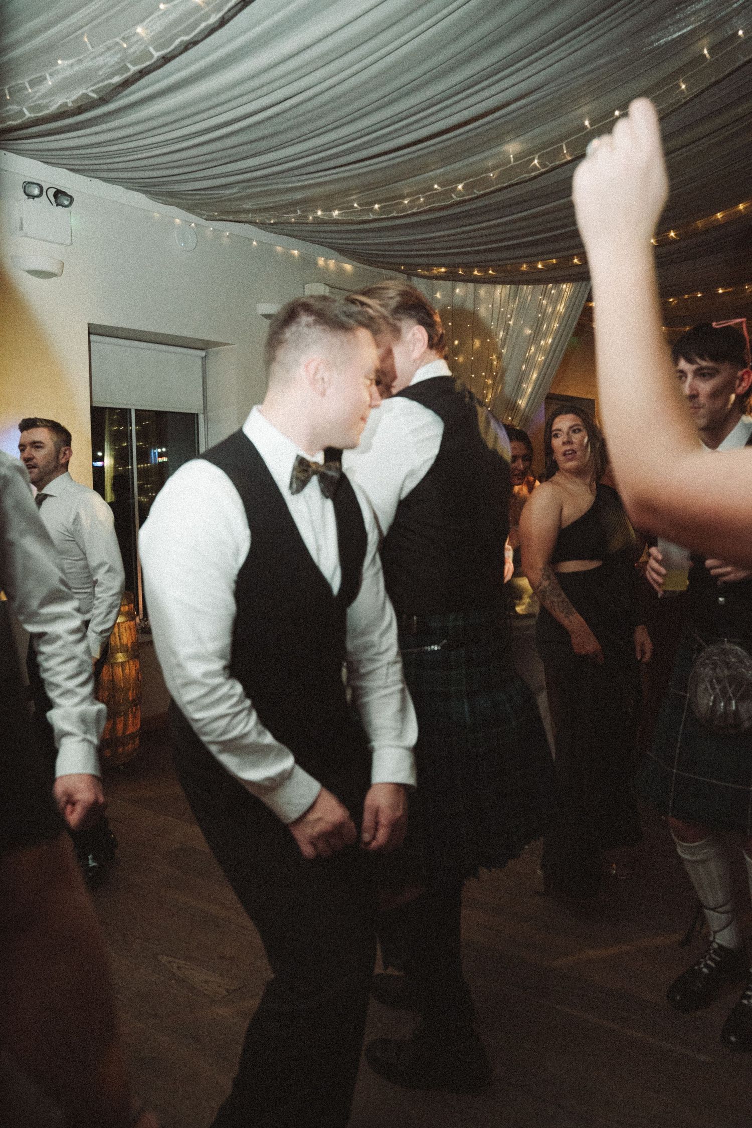 Wedding guests dancing on a wooden floor under draped fabric and string lights.