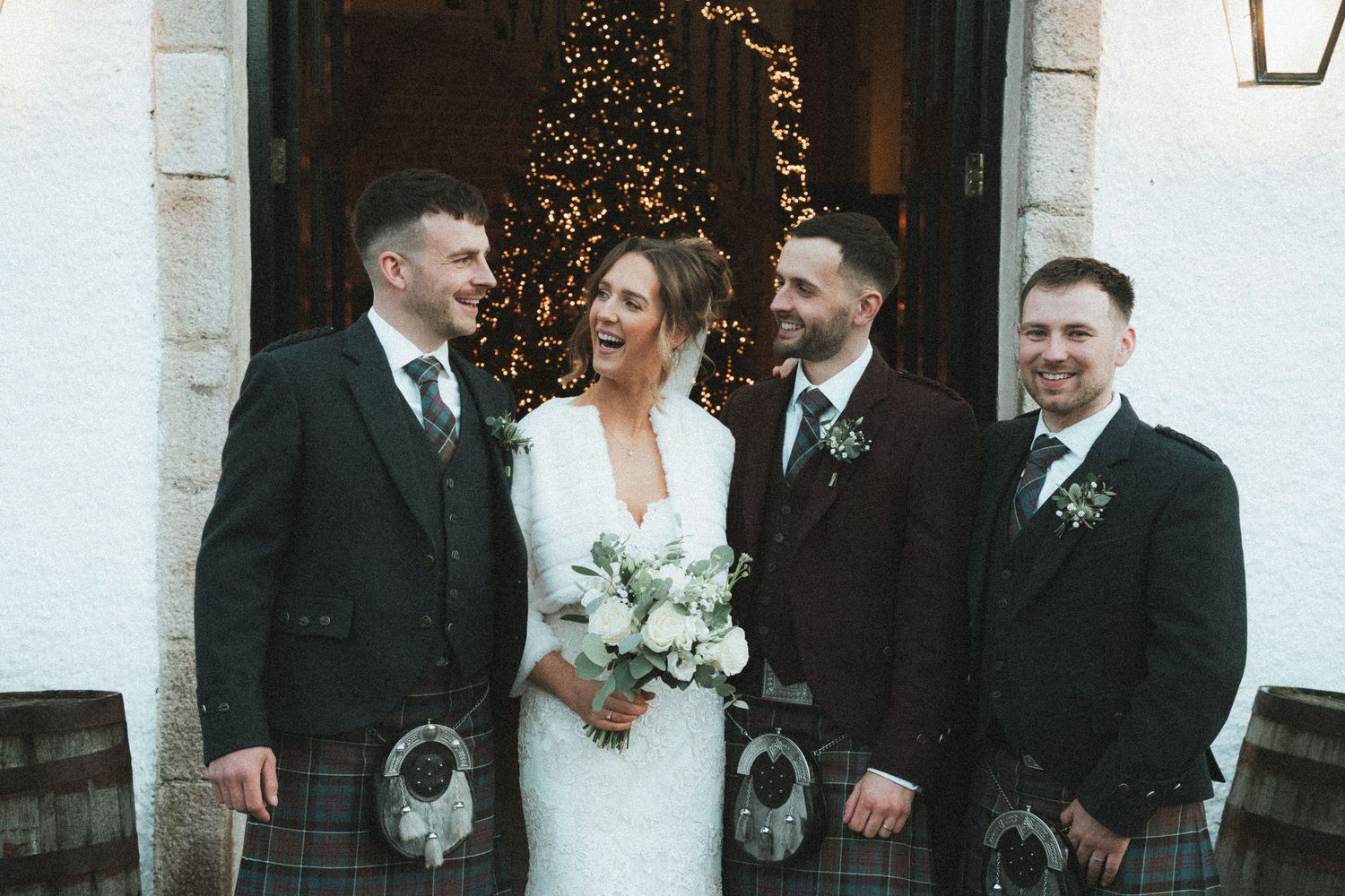 Wedding party in traditional Scottish attire poses in front of church entrance with Christmas lights.