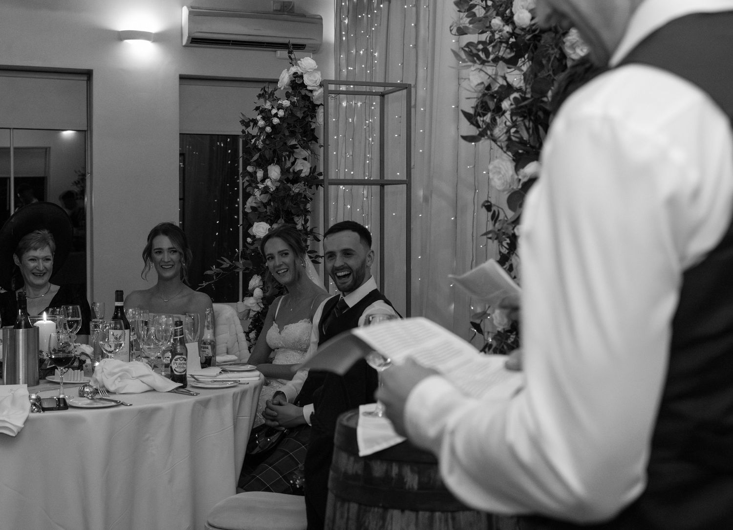Black and white photo of wedding reception guests seated at decorated tables during speeches.