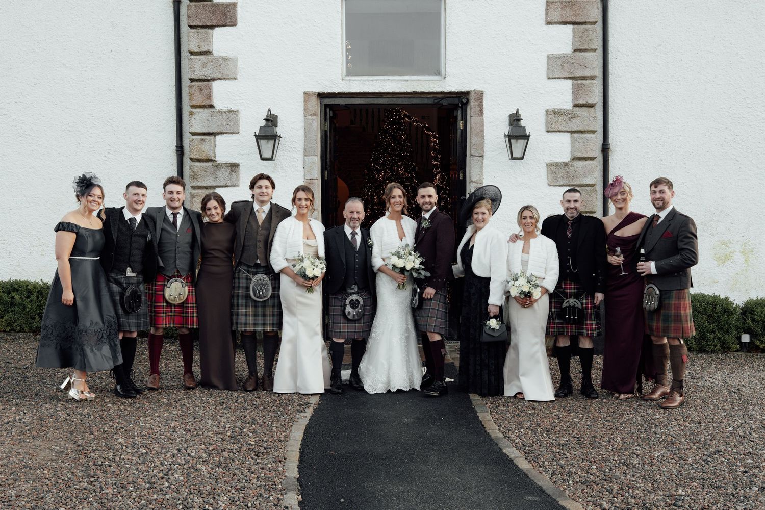 Wedding group poses in front of white stone building entrance with traditional Scottish kilts and formal attire.