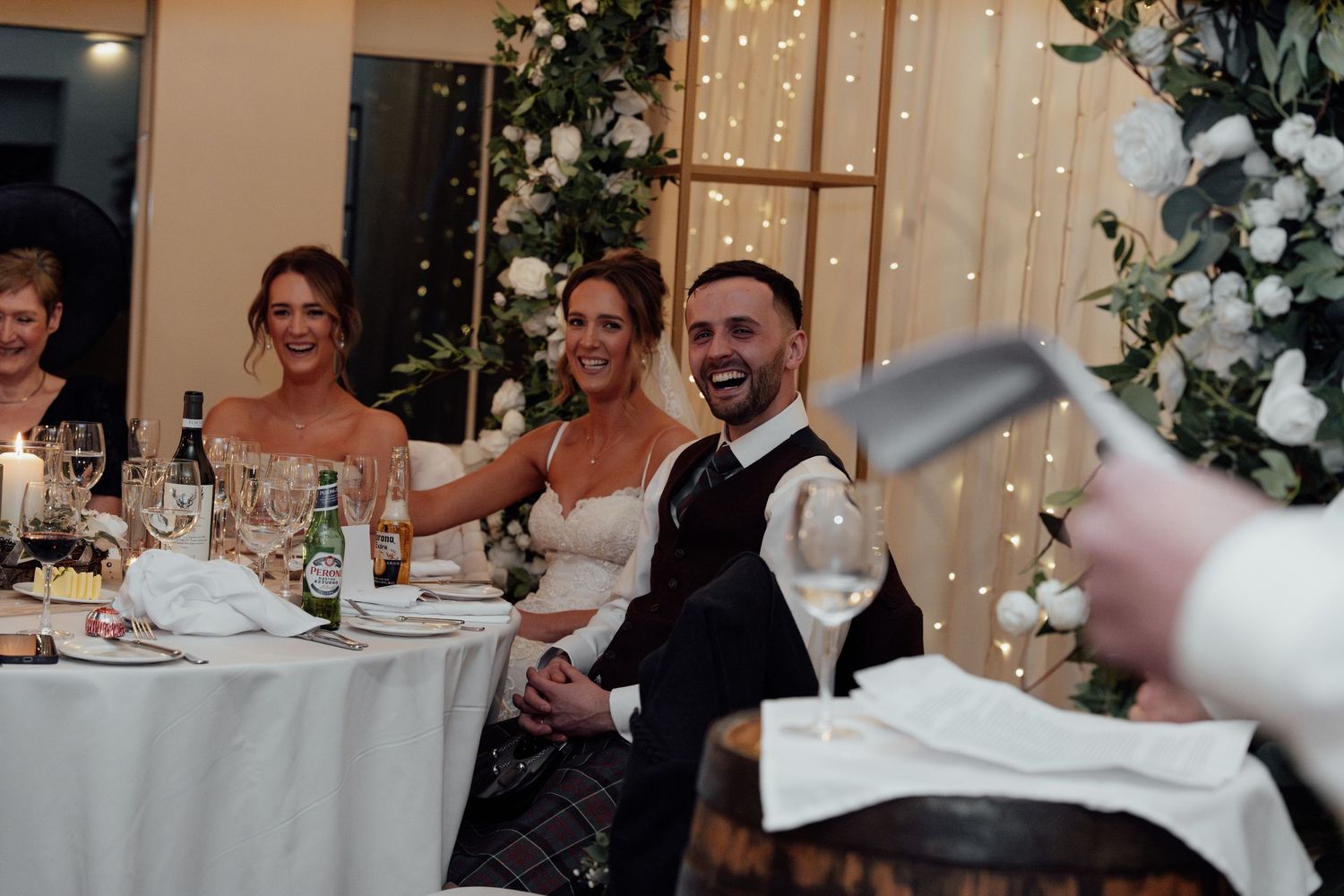 Wedding party seated at decorated tables with fairy light backdrop and floral arrangements.