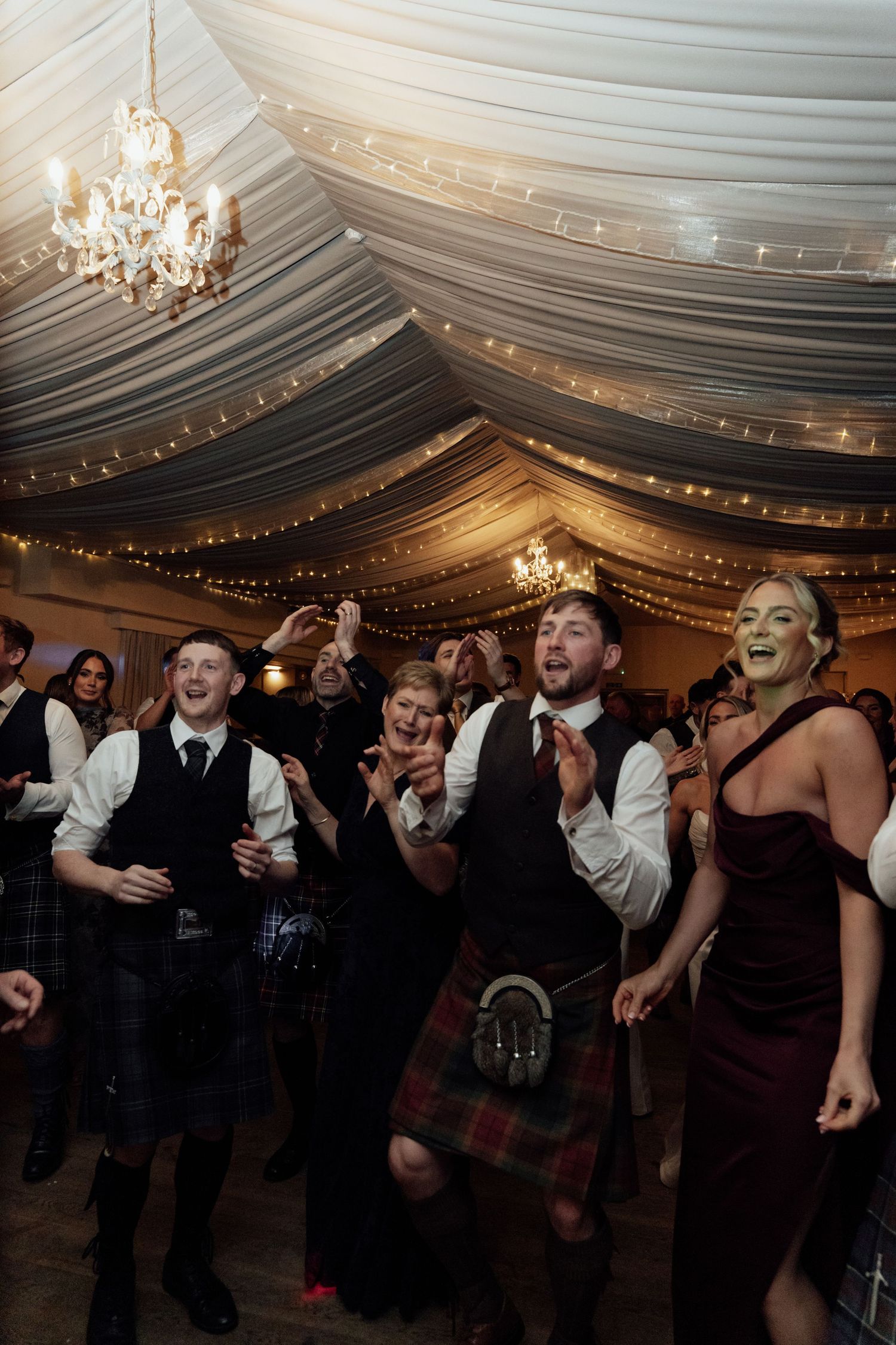 Wedding guests dancing and celebrating under an elegant draped ceiling with chandeliers.