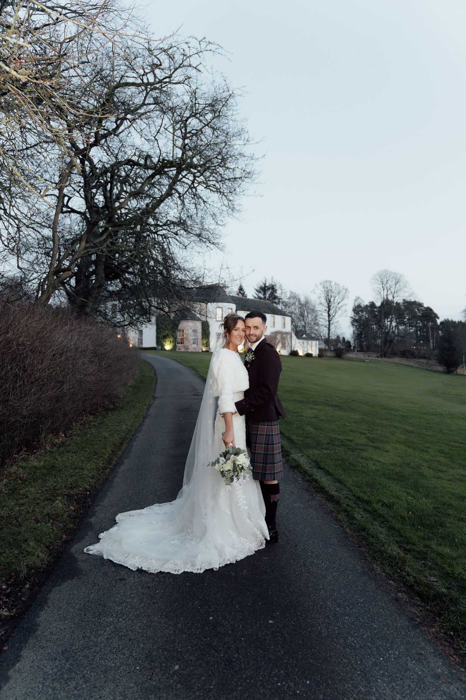 Newlyweds pose on a winding path with a beautiful white manor house in the background.