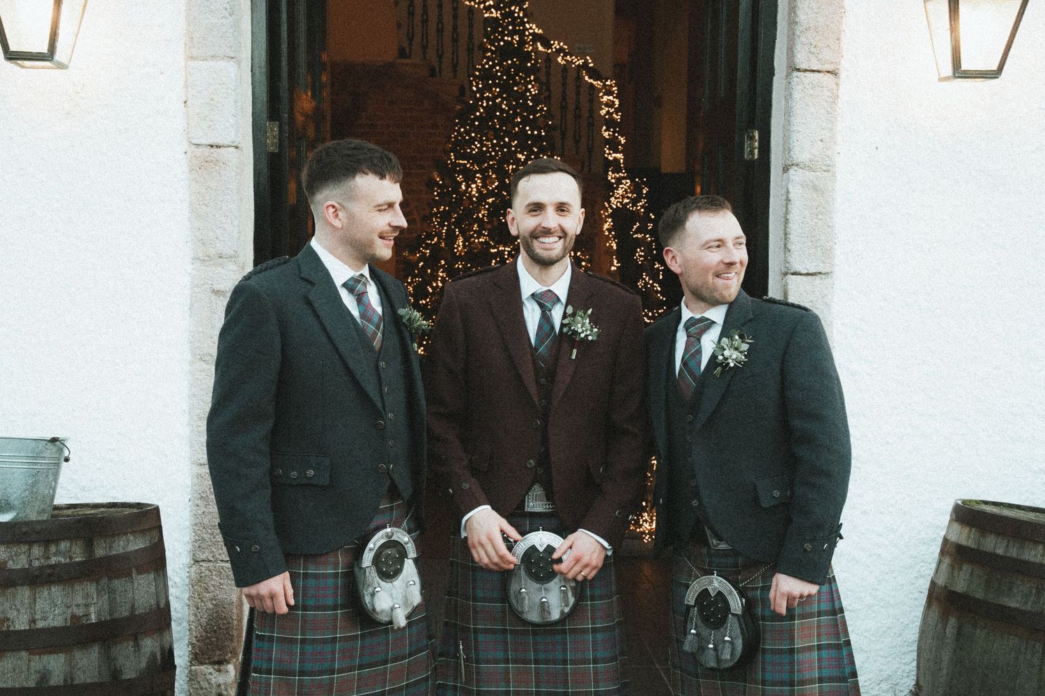 Three groomsmen in traditional Scottish kilts pose in front of decorated entrance.