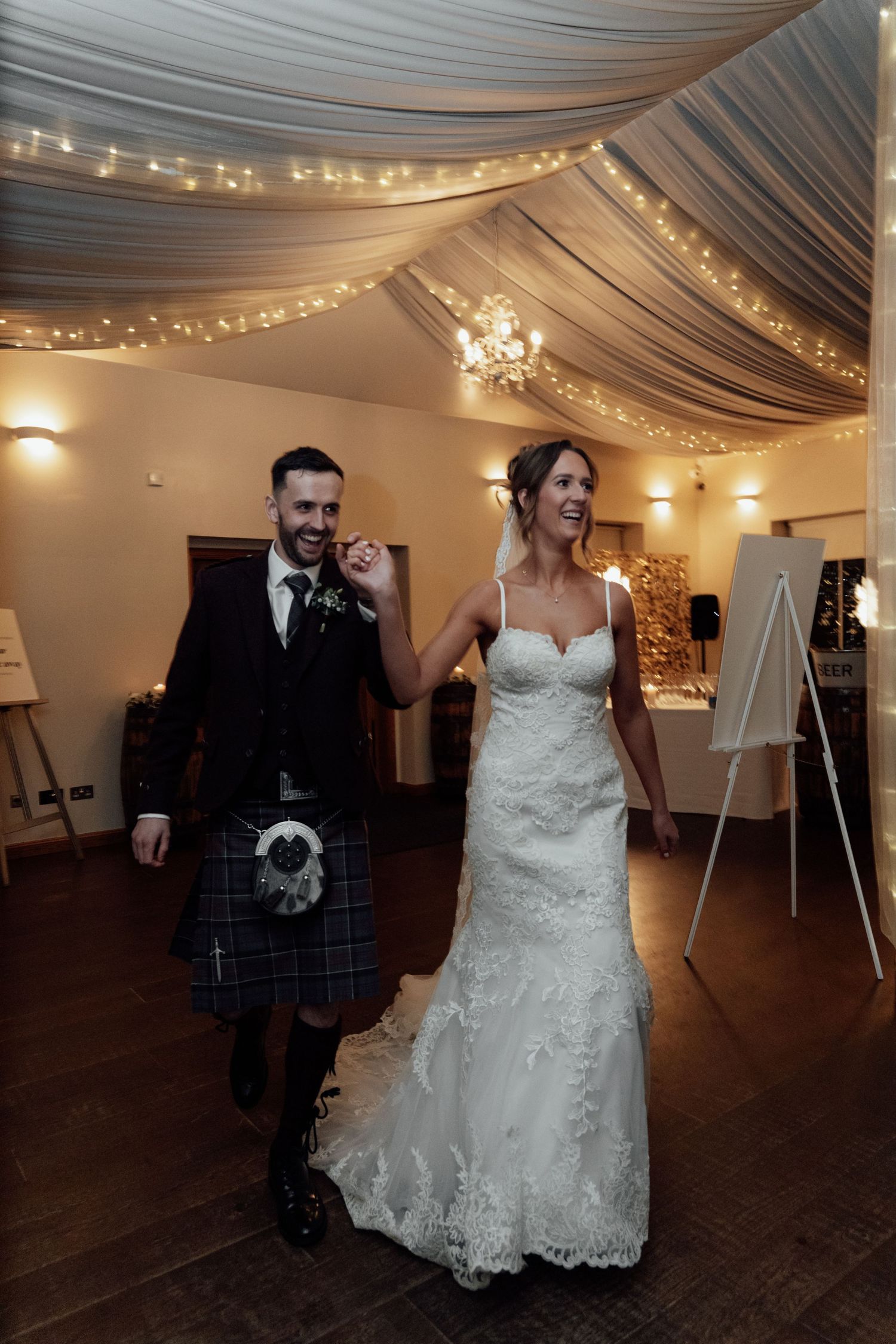 Wedding couple walks under draped fabric ceiling with twinkling lights at reception venue.