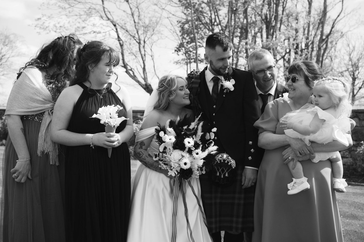Black and white wedding photo of bride, groom in kilt, and family group smiling outdoors.