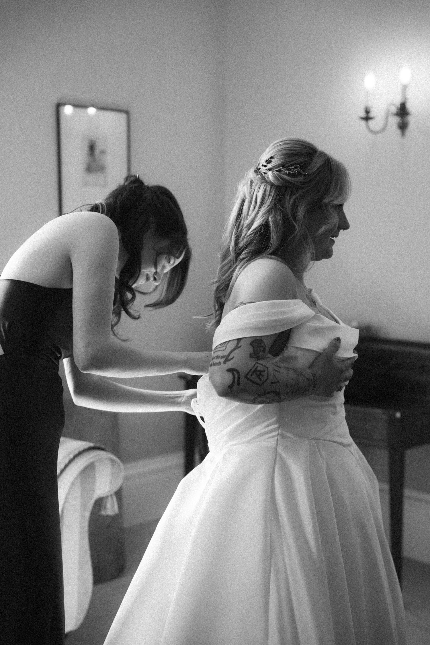 Bridesmaid helping bride into white off-shoulder wedding gown in black and white photo.