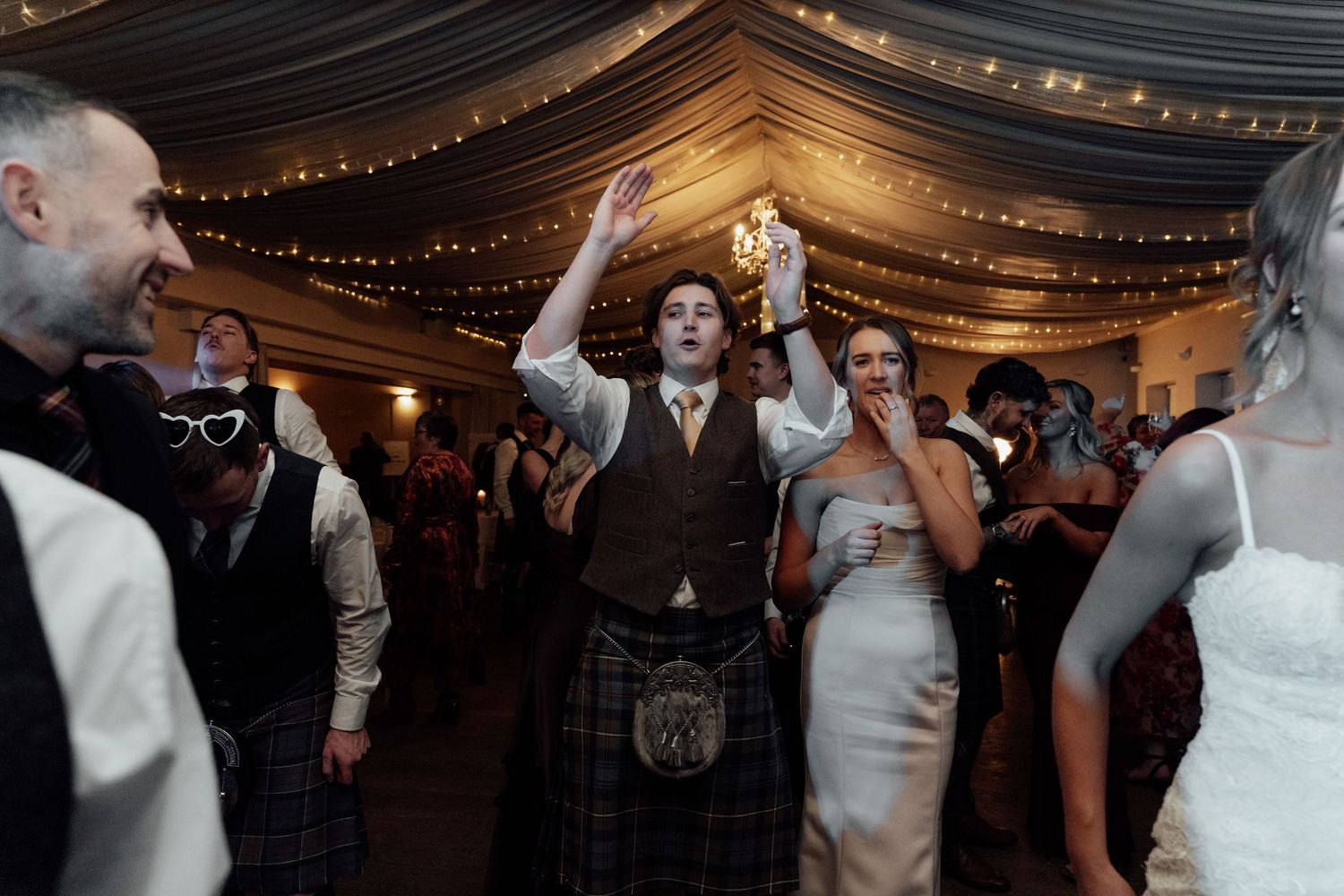Energetic dancing at wedding reception with dark draped ceiling and string lighting.
