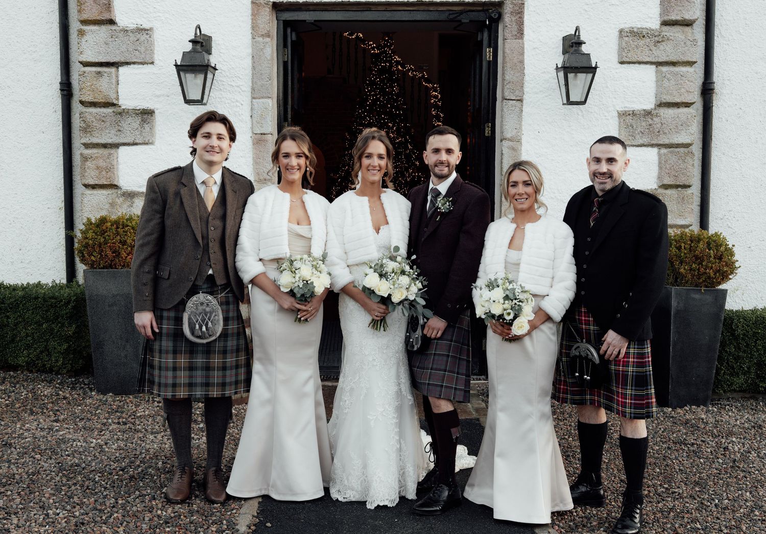 Wedding party in traditional Scottish and white formal attire poses together outside venue entrance.