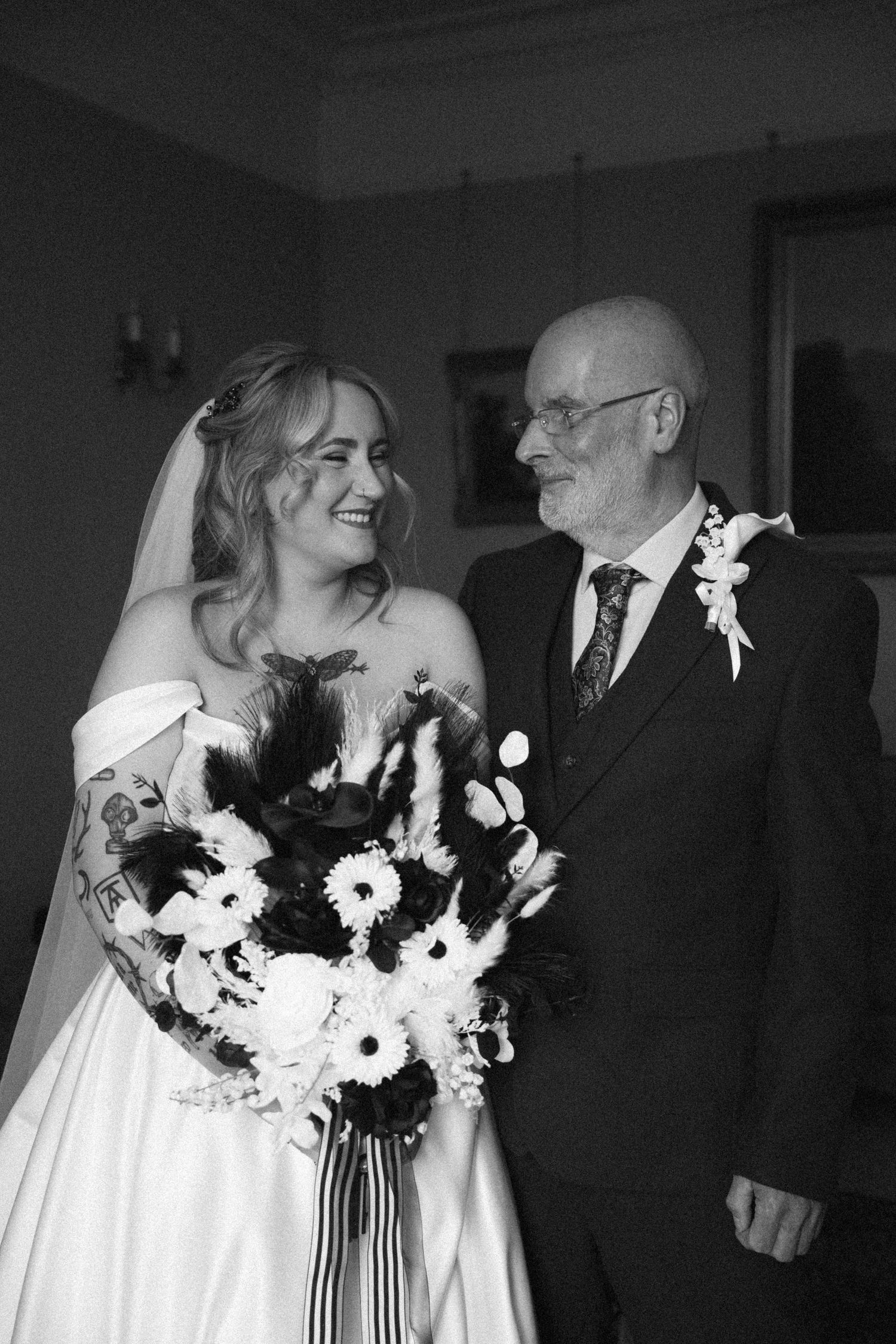 Black and white photo of smiling bride with tattoos and bouquet standing with older man in suit.