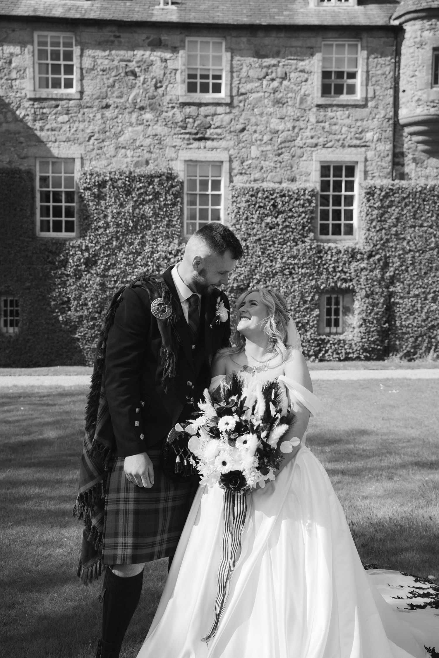 Black and white photo of a groom in a kilt and bride in a white gown embracing outside a stone castle.