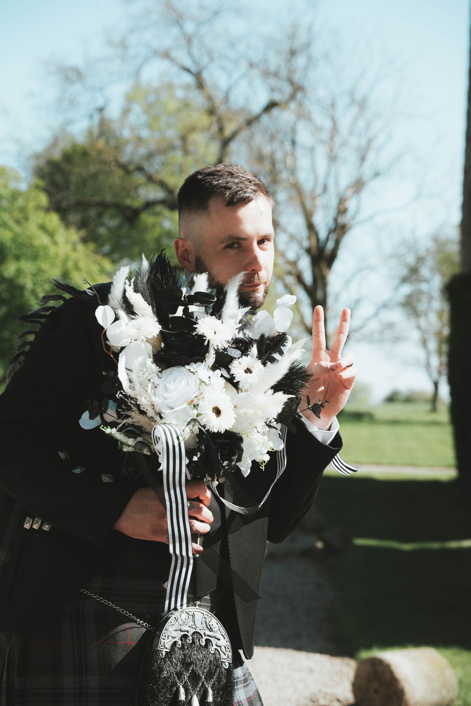 Groom in Scottish kilt holding black and white bouquet making peace sign outdoors on sunny day.