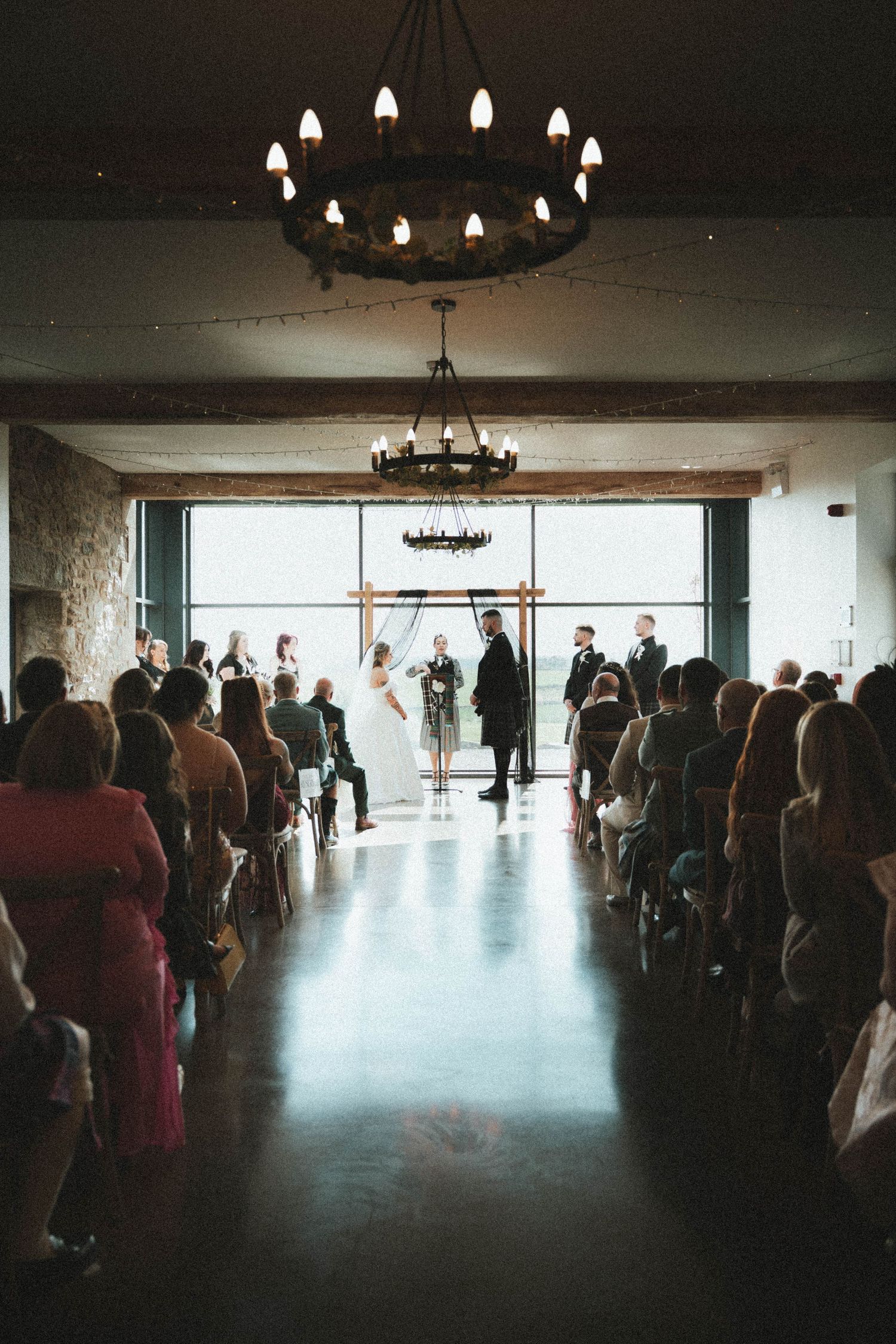 Wedding ceremony in rustic indoor venue with chandelier lighting, guests seated in rows as couple exchanges vows at altar.