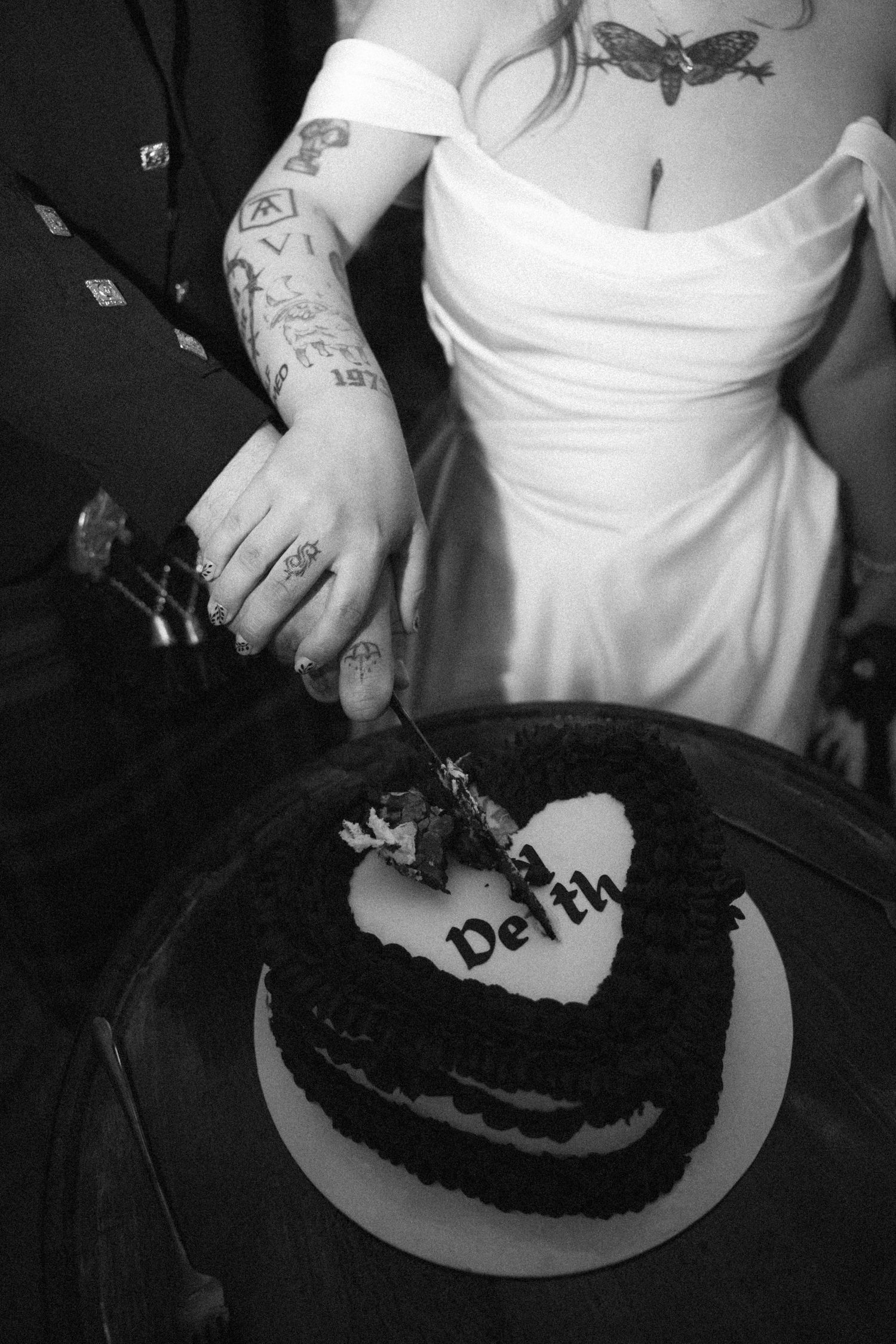 Tattooed couple cutting heart-shaped black wedding cake with Death inscription in black and white.