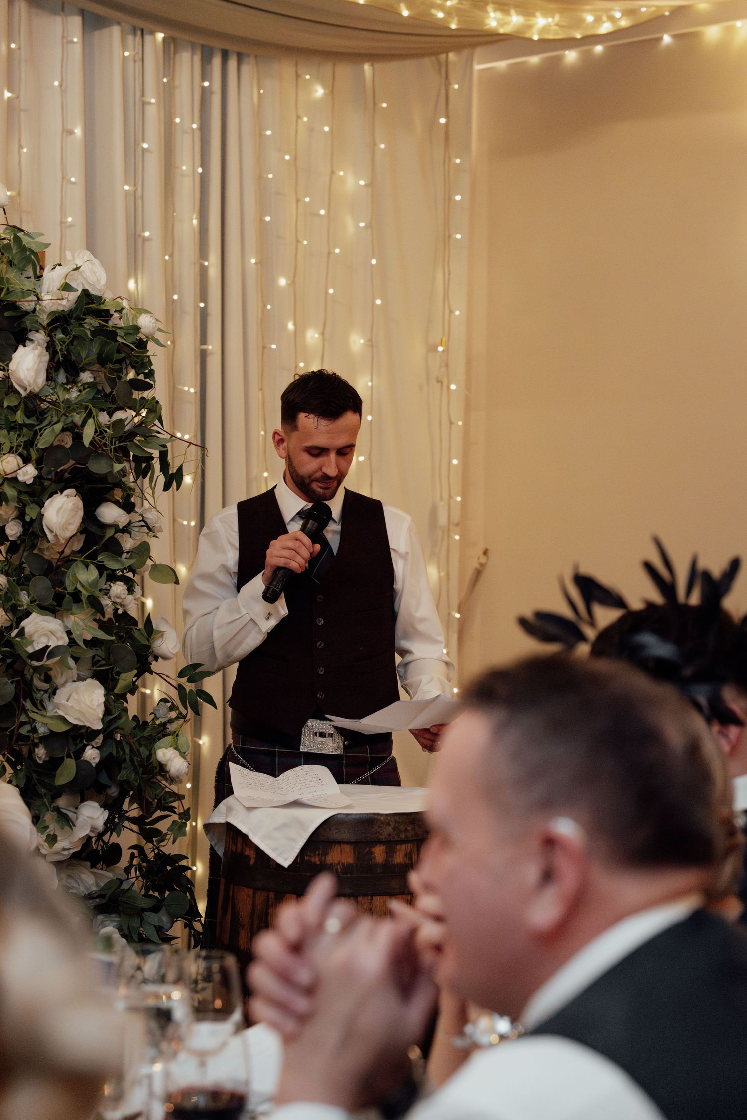Wedding speeches being given during reception dinner with decorative lights and greenery.