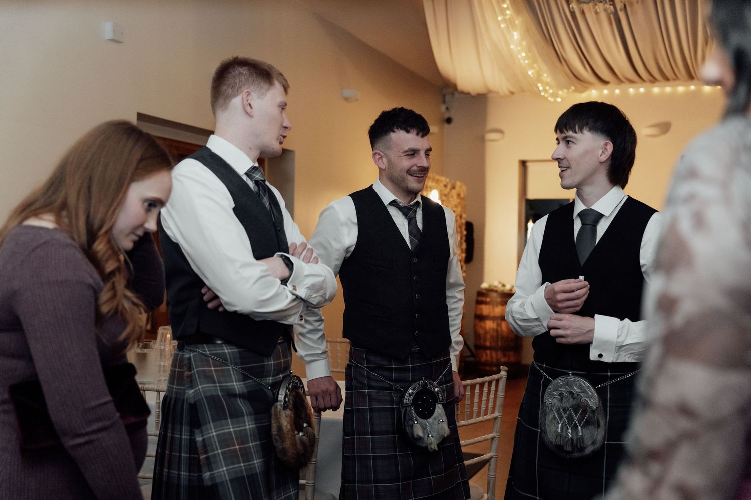 Wedding party members in formal wear and kilts gather during the reception.