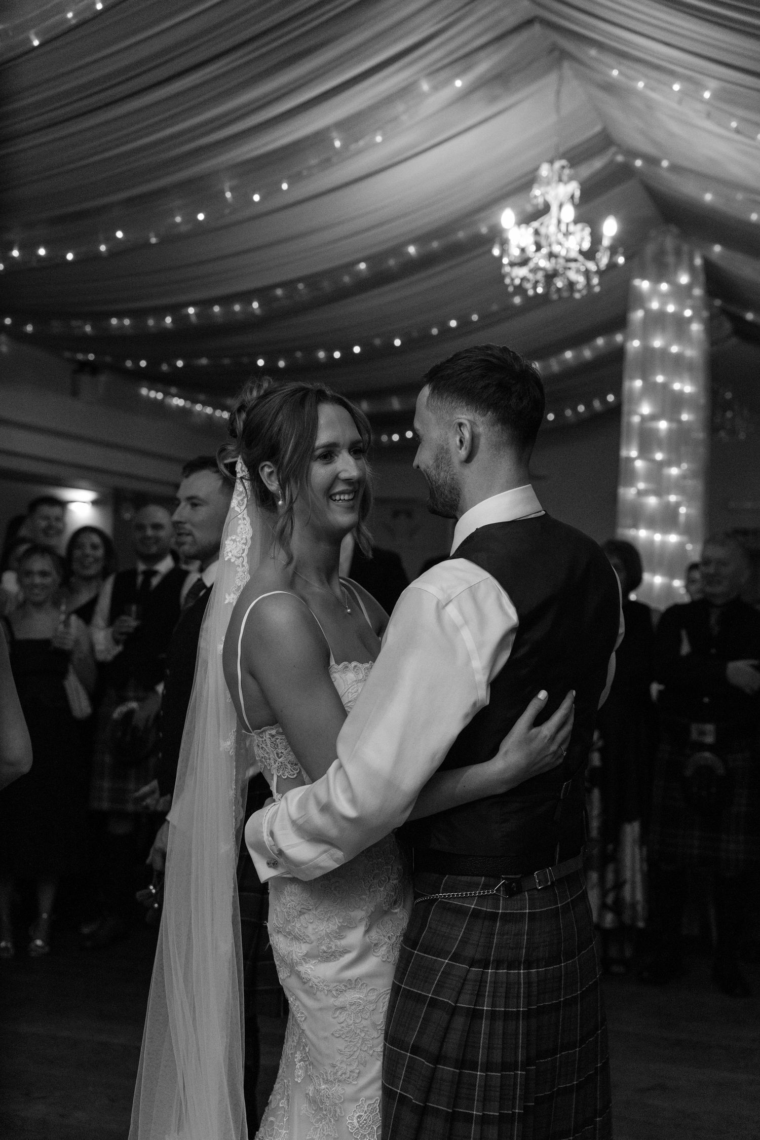 Wedding couple embraces during romantic dance under twinkling reception tent lights.
