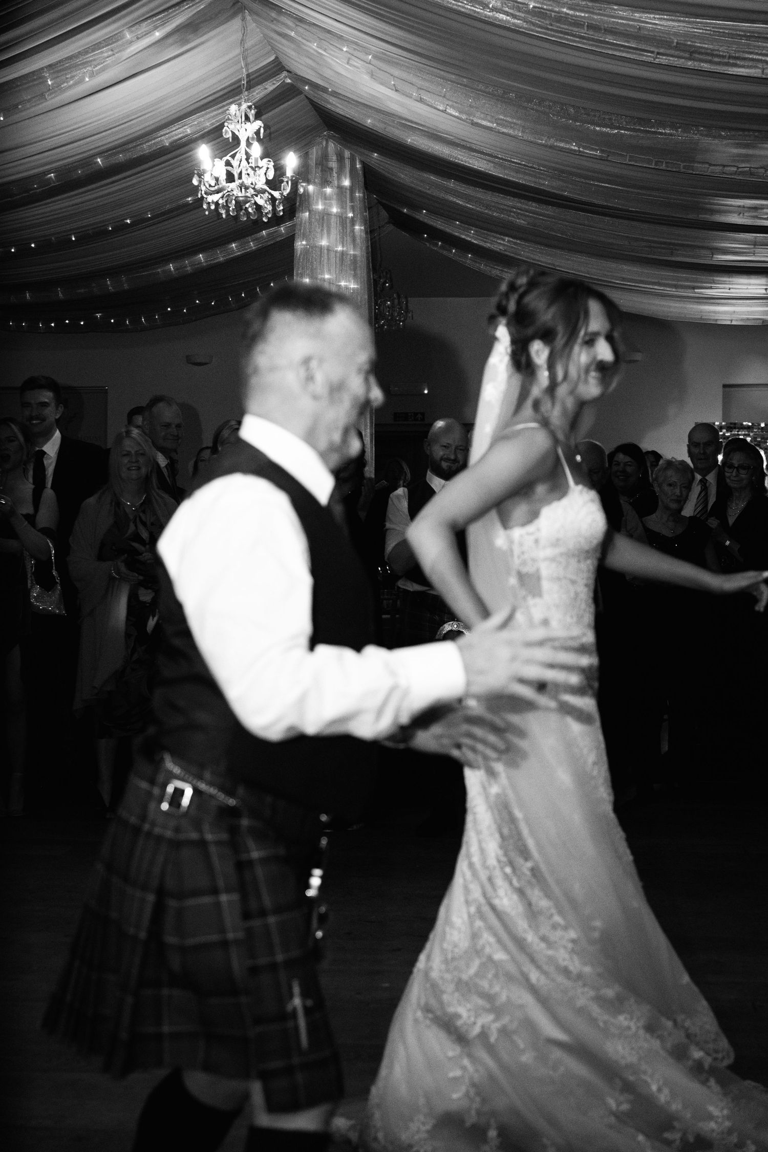 Wedding couple dances in an elegant venue decorated with fabric draping and chandeliers.