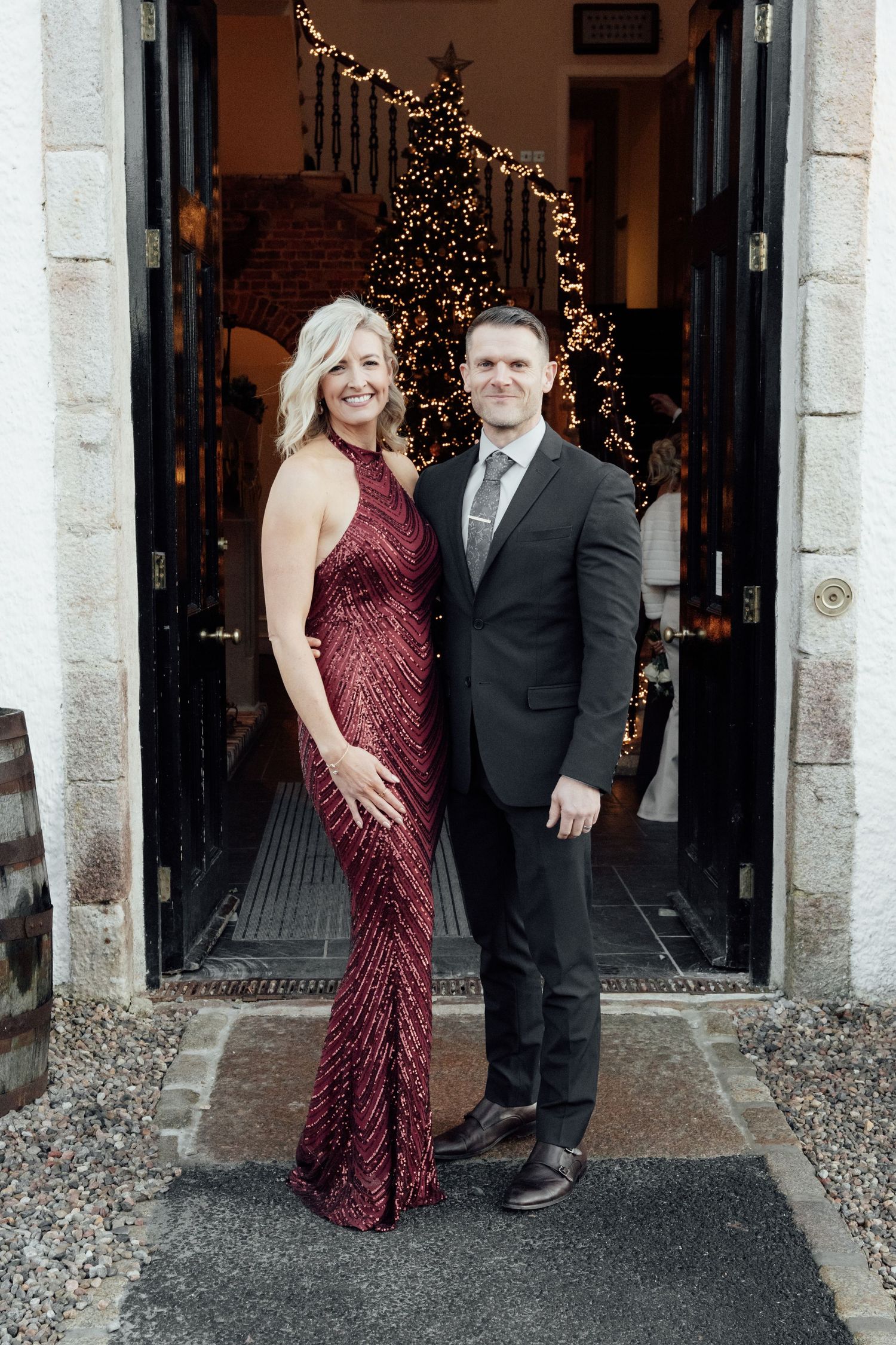 A couple poses by a doorway, with the woman wearing a sparkly burgundy gown and man in a black suit.