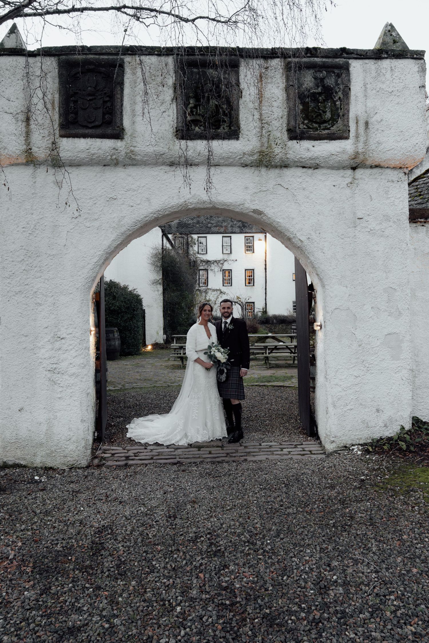 Wedding couple standing under white archway in historic courtyard setting.