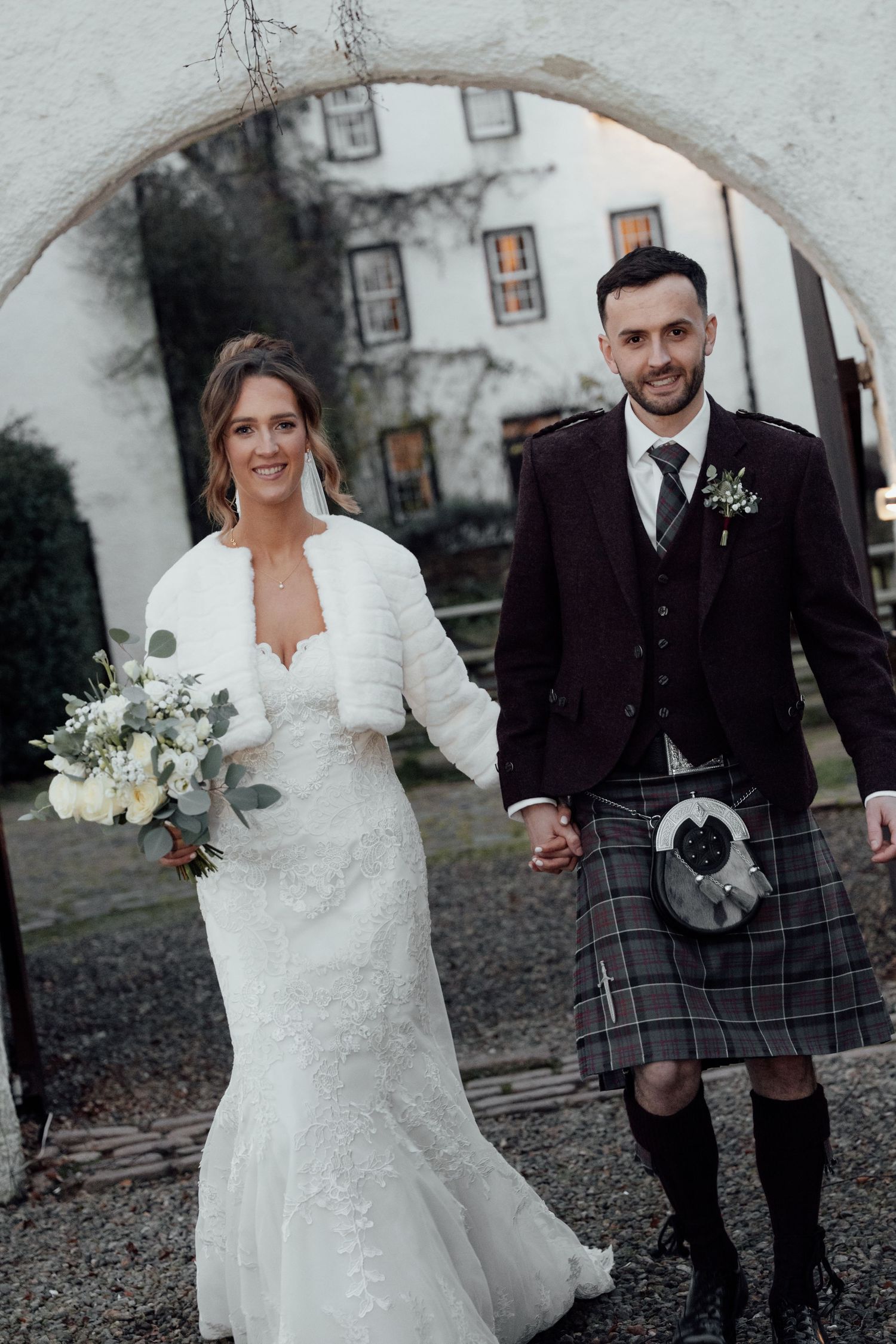 Wedding couple poses under stone arch, bride in white dress with fur wrap and groom in burgundy jacket and kilt.