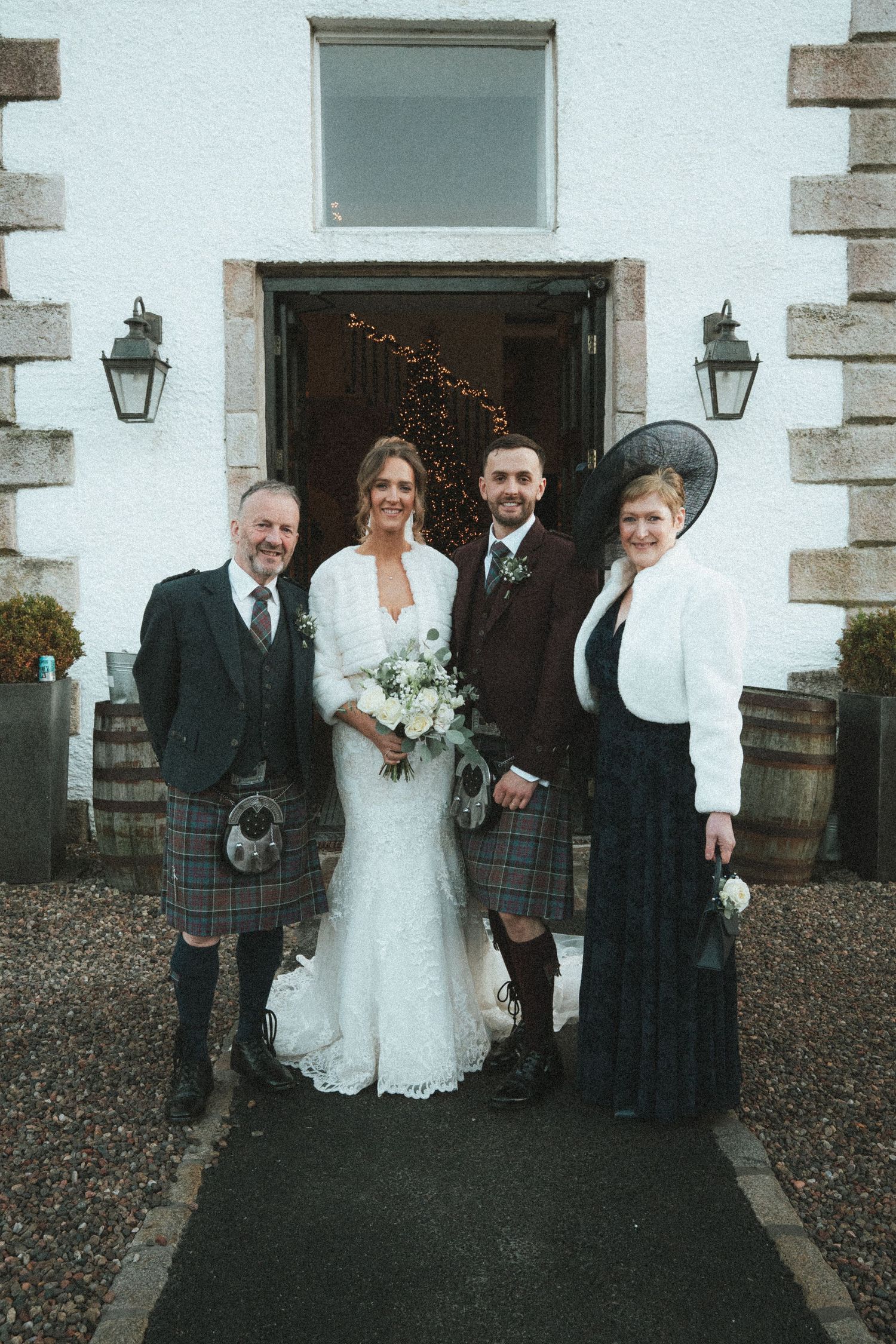 A formal wedding group photo with Scottish kilts and traditional attire in front of a white building.