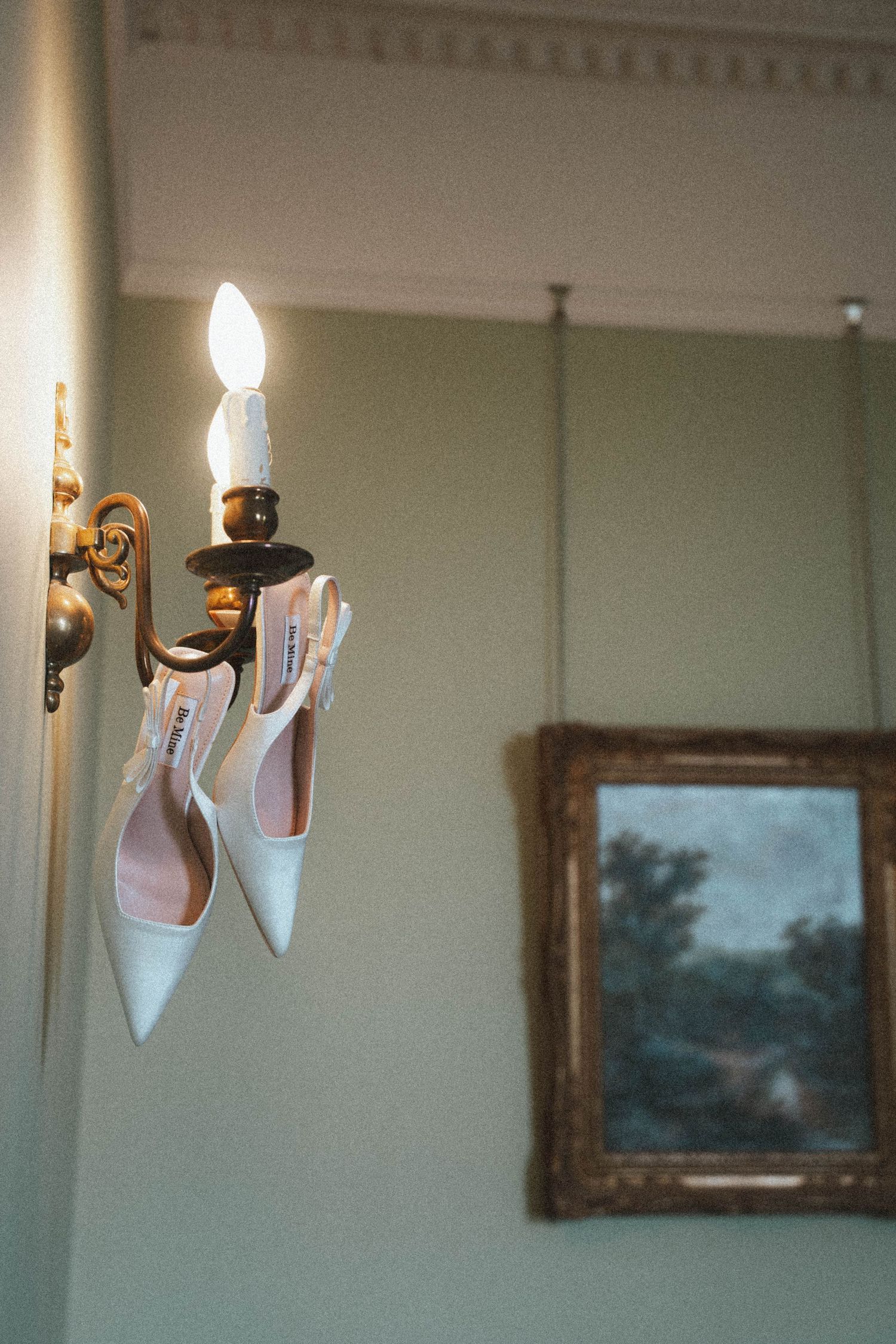 White bridal heels hanging on a gold wall sconce light fixture in an elegant room with a framed painting.