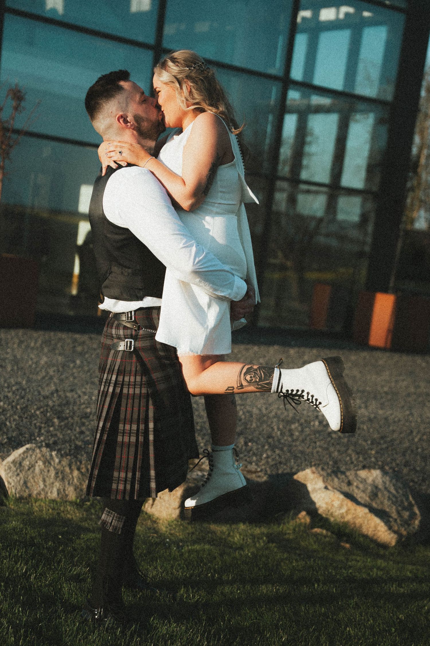 Groom in tartan kilt lifts bride in white mini dress and white Dr. Martens boots outdoors at sunset.