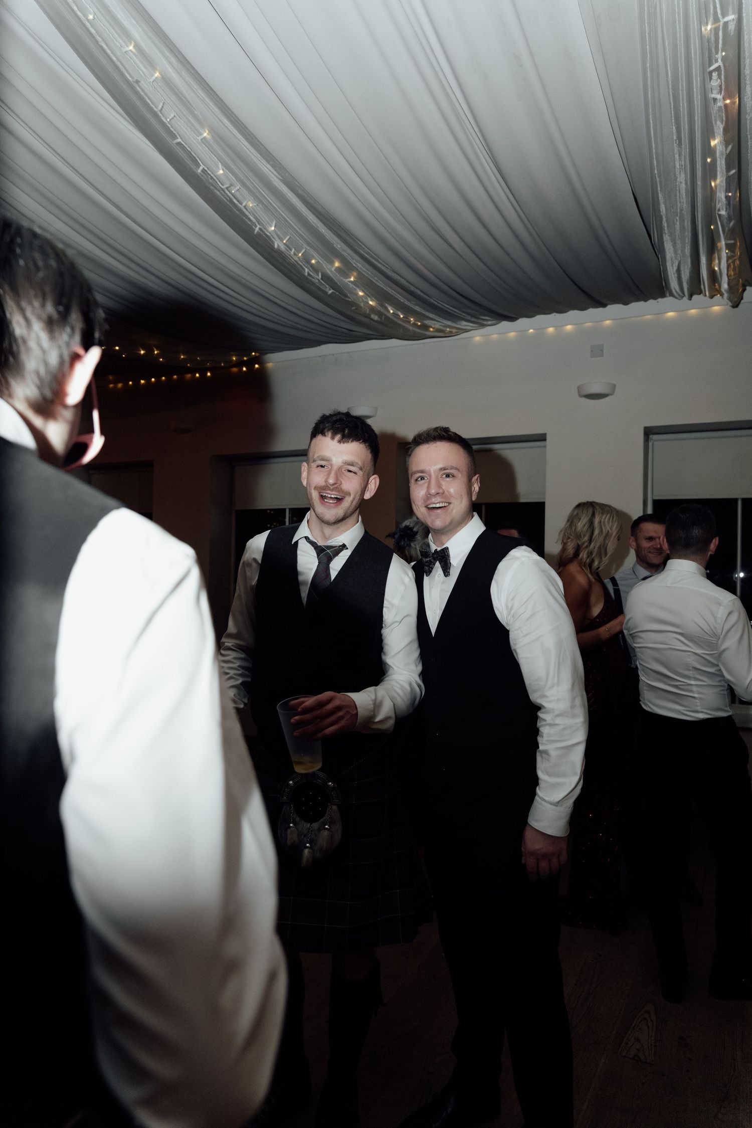 Wedding guests in formal attire share laughs during a reception under draped fabric ceiling with string lights.