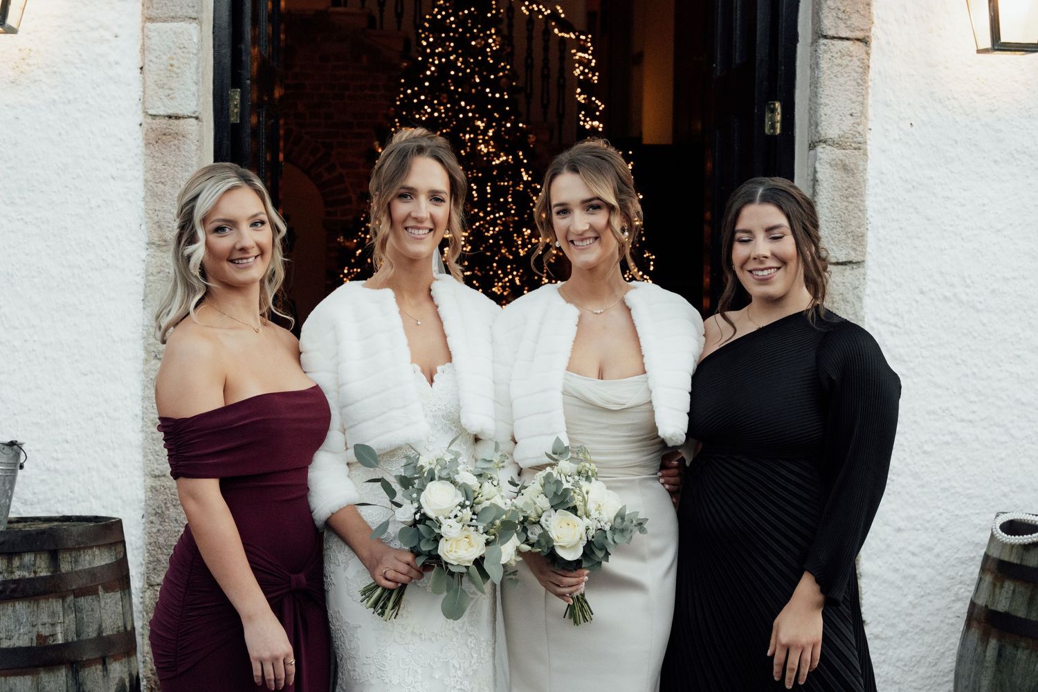 Wedding party members smile together outside venue entrance with holiday lights visible.