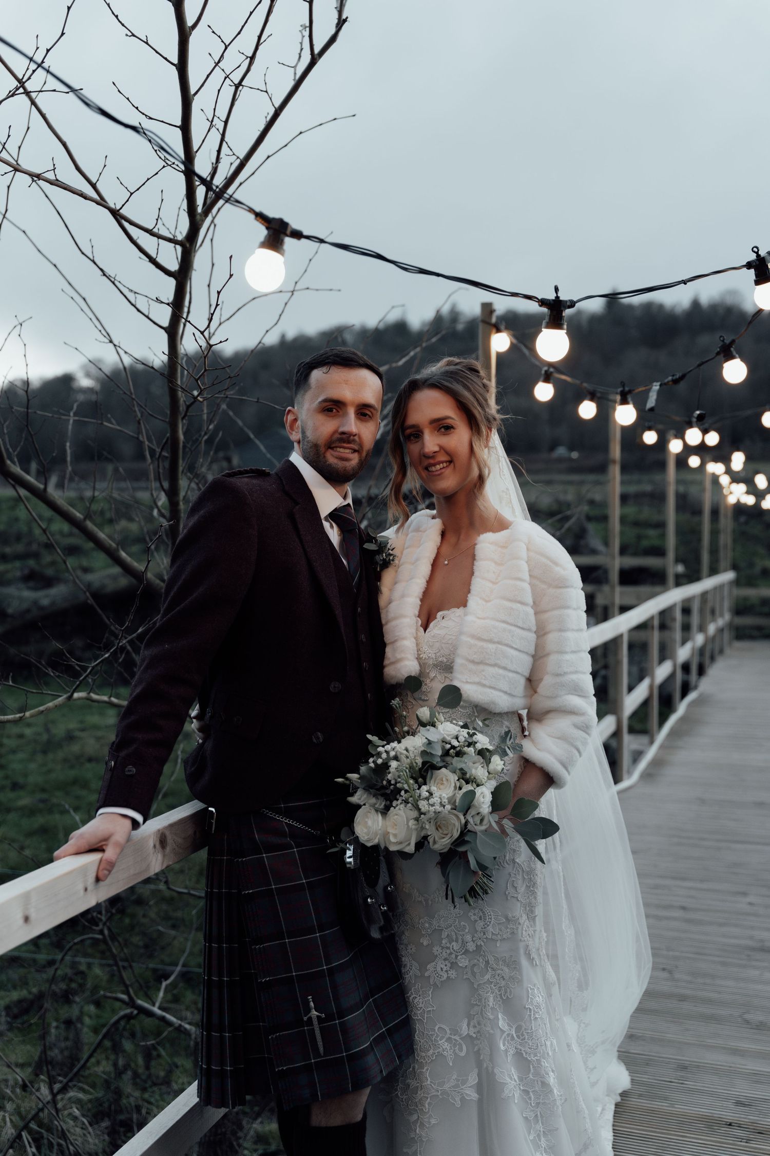 A romantic wedding moment on a wooden bridge decorated with string lights at dusk.