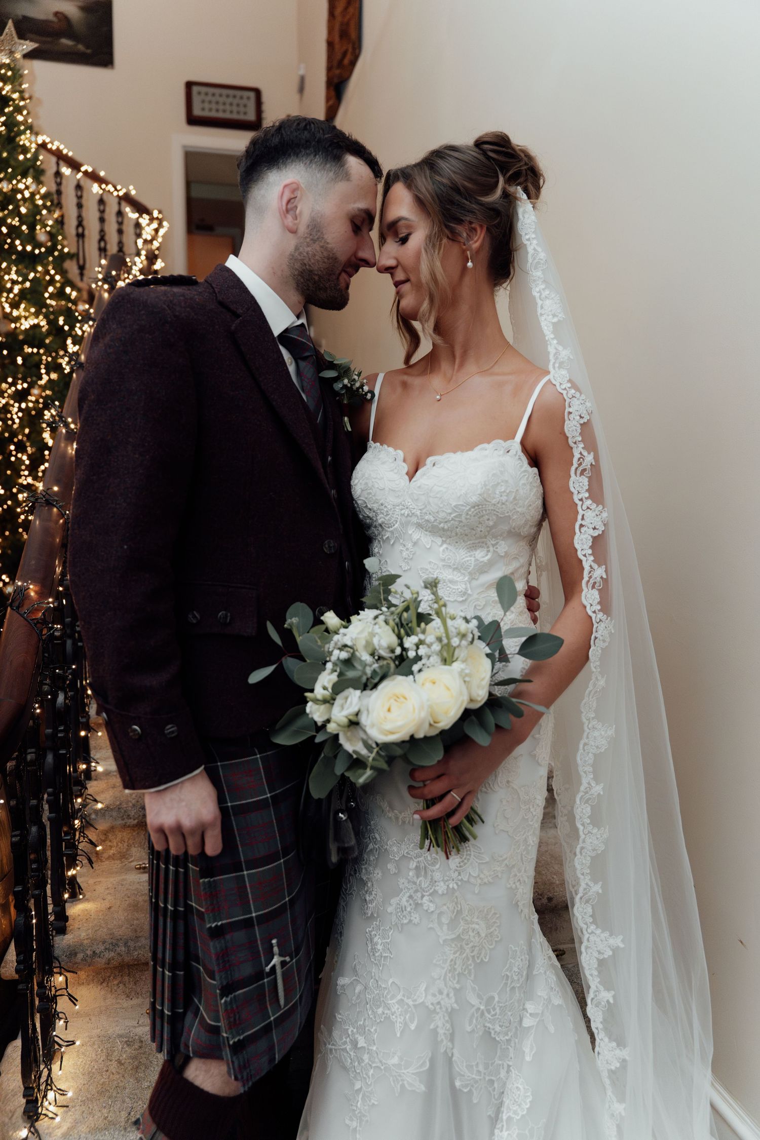 Elegant wedding portrait featuring Scottish kilt and lace bridal gown against lit Christmas tree.
