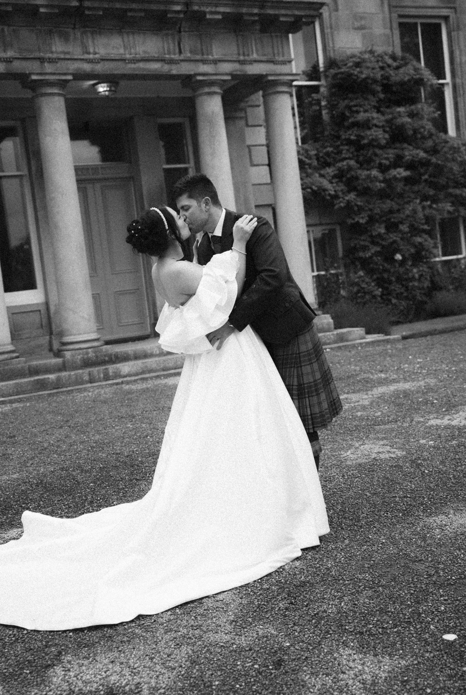 Black and white photo of a bride and groom sharing a romantic kiss outside a grand building with columns.