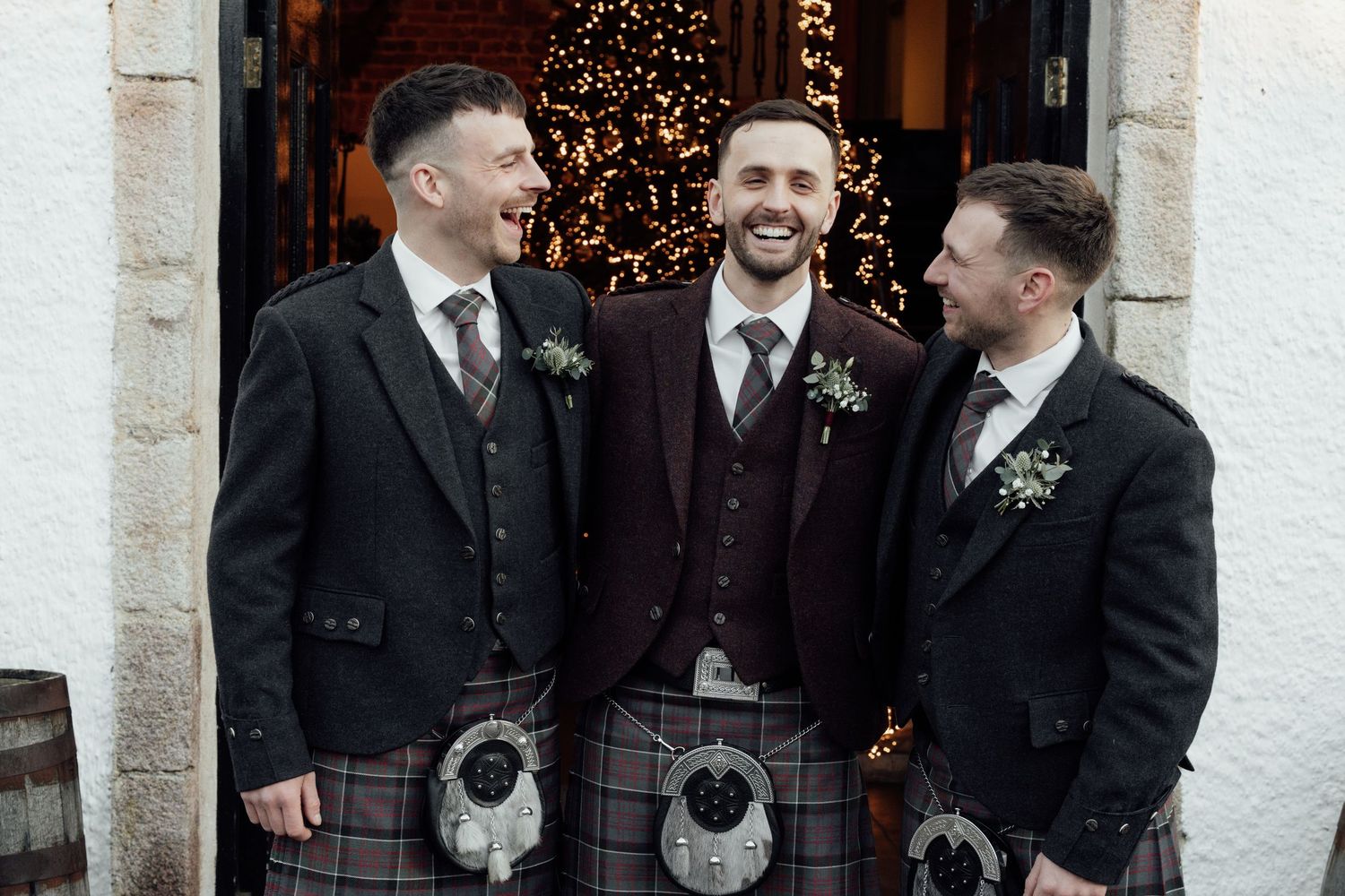 Three groomsmen in Scottish formal wear share a candid moment at decorated venue entrance.