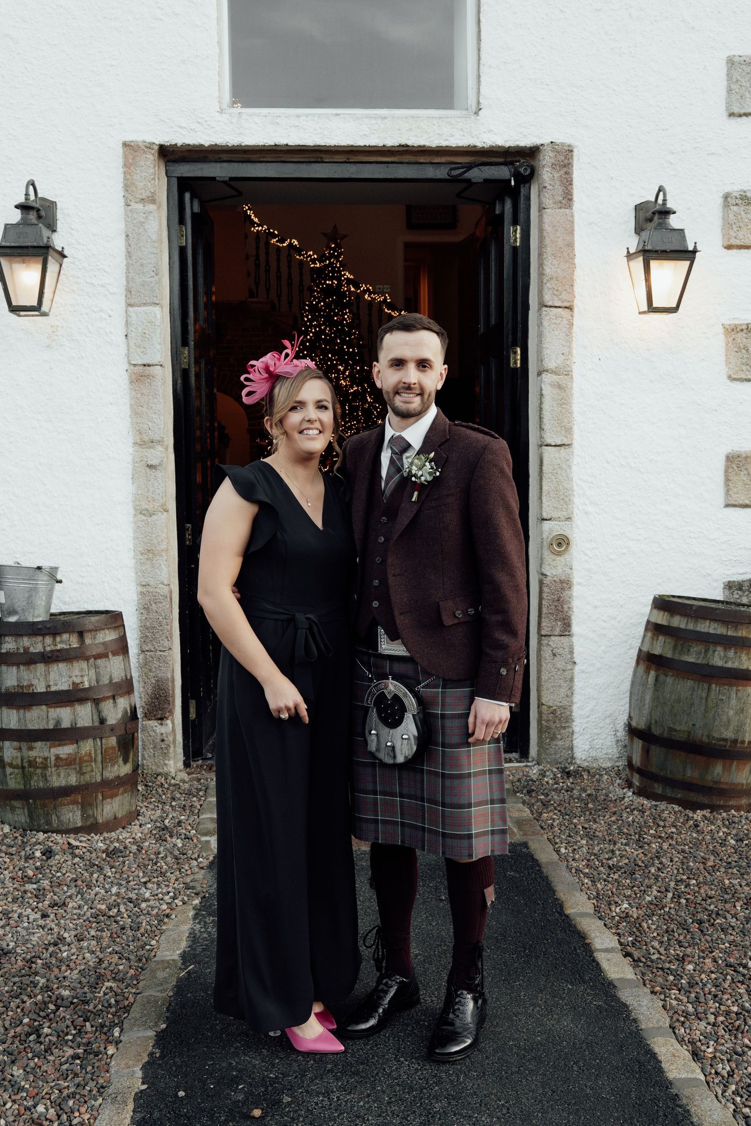 Wedding guests in formal attire pose against white stone wall with wooden barrels and lanterns.