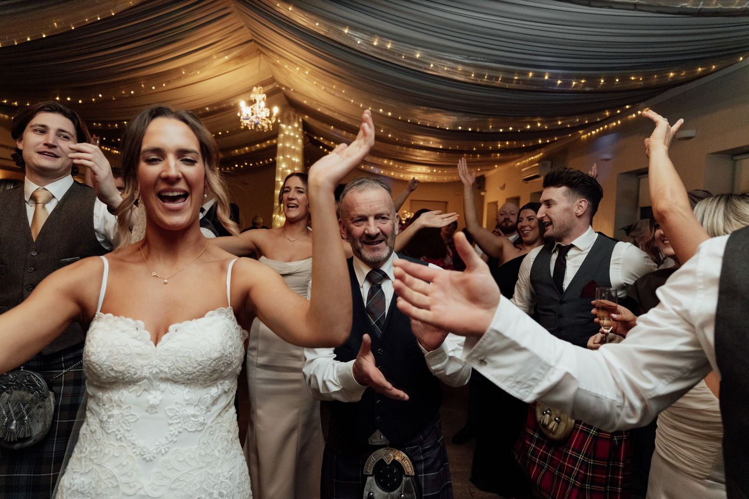 Wedding party dancing and celebrating enthusiastically under decorated reception tent.