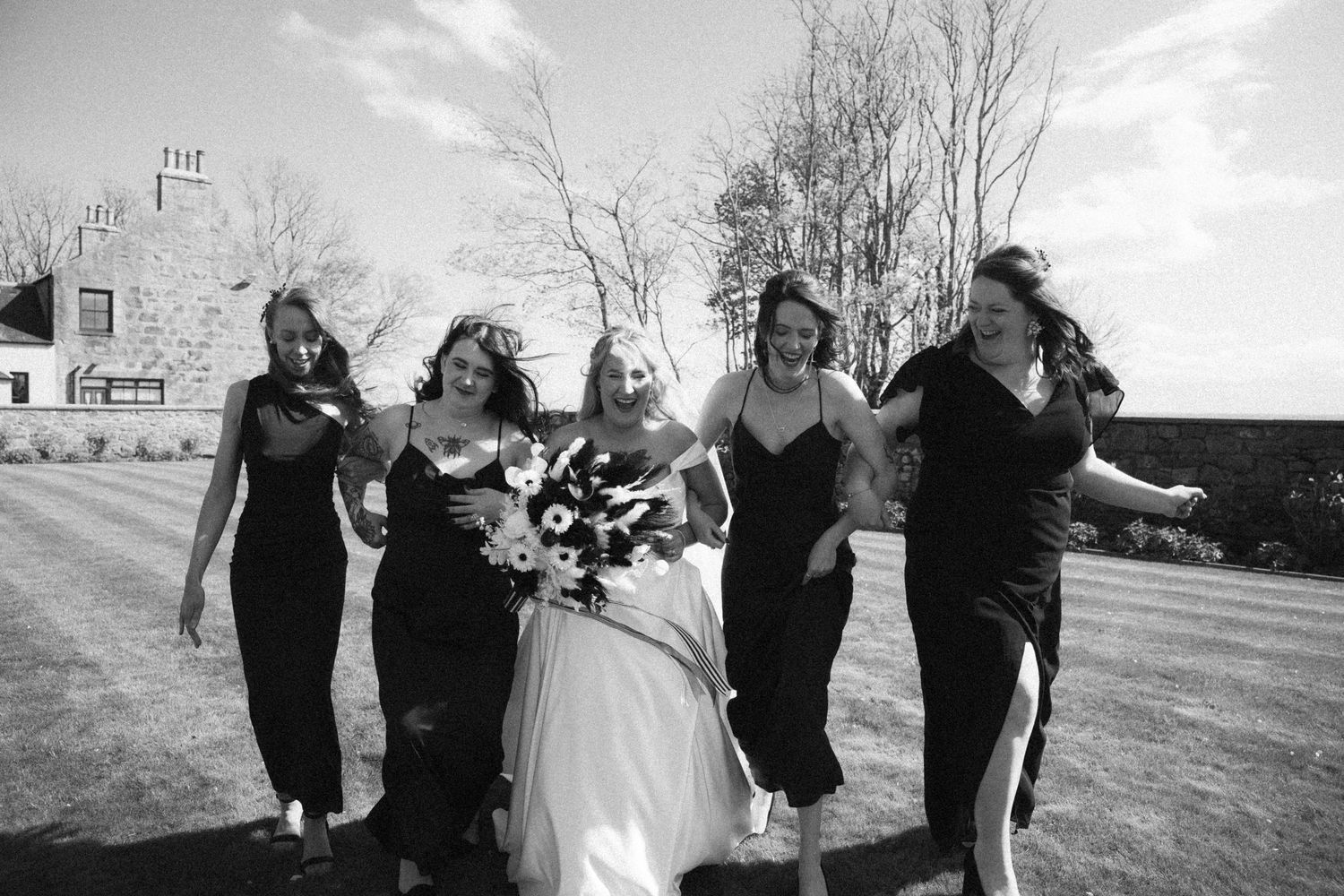 Black and white photo of bride and bridesmaids in black dresses laughing outdoors on wedding day.