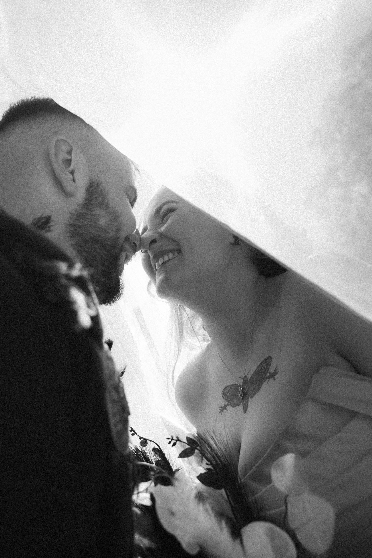 Intimate black and white under-veil photo of bearded groom kissing smiling tattooed bride at their wedding ceremony.